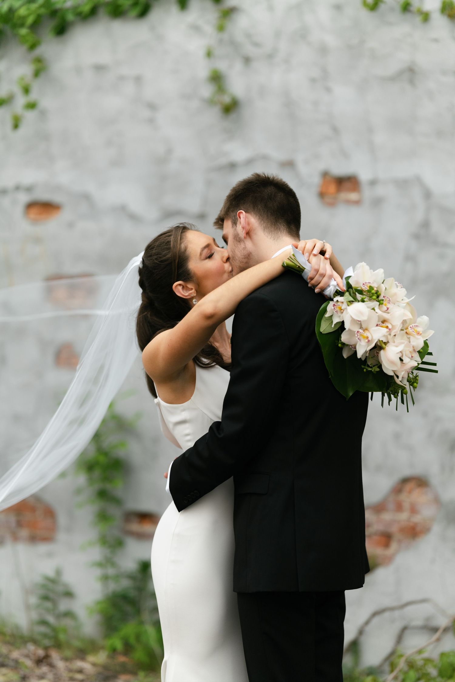 Intimate moment between newlyweds sharing embrace against textured wall with trailing veil.
