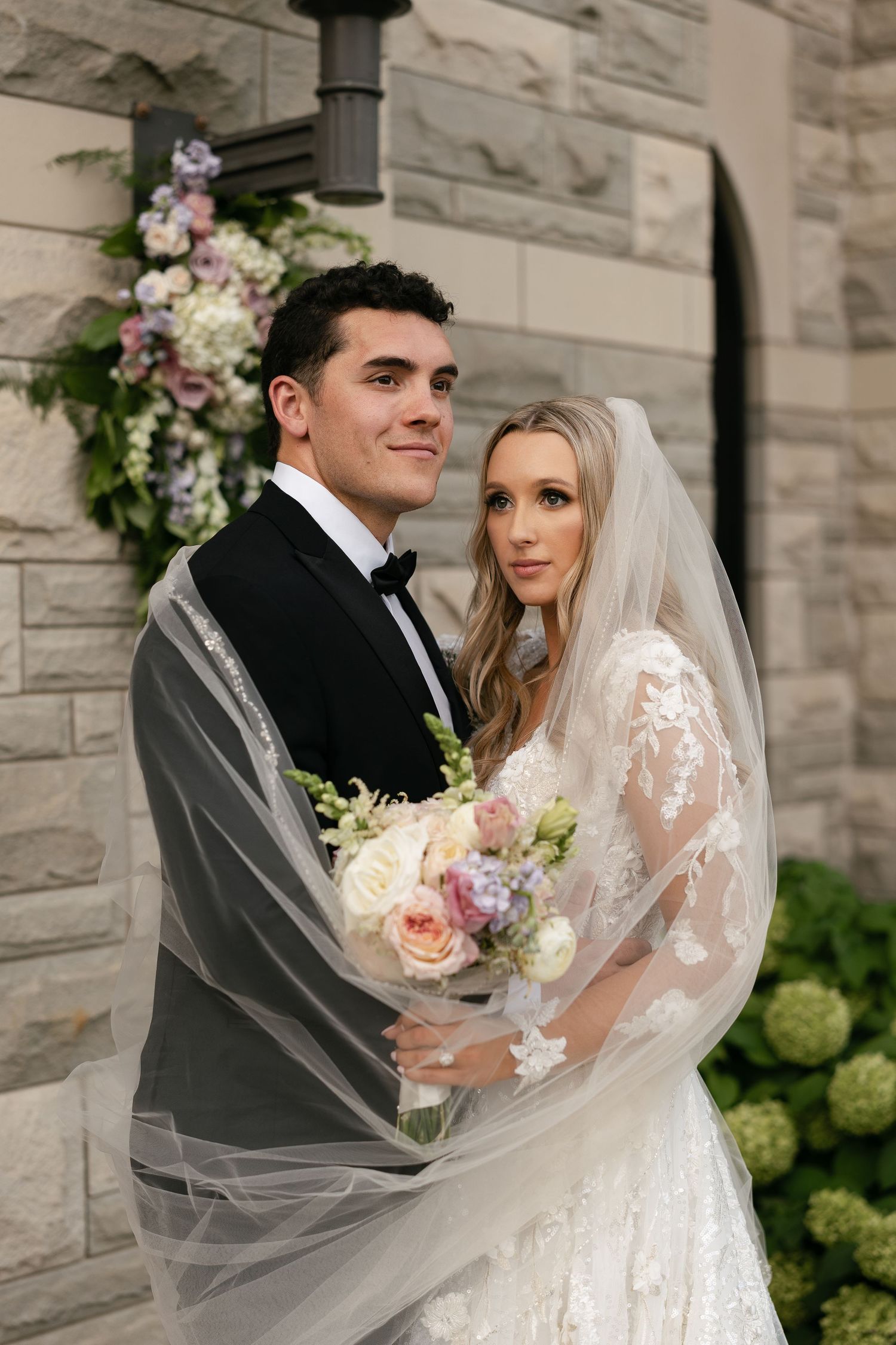 A romantic wedding portrait with soft florals and flowing veil against a stone building backdrop.