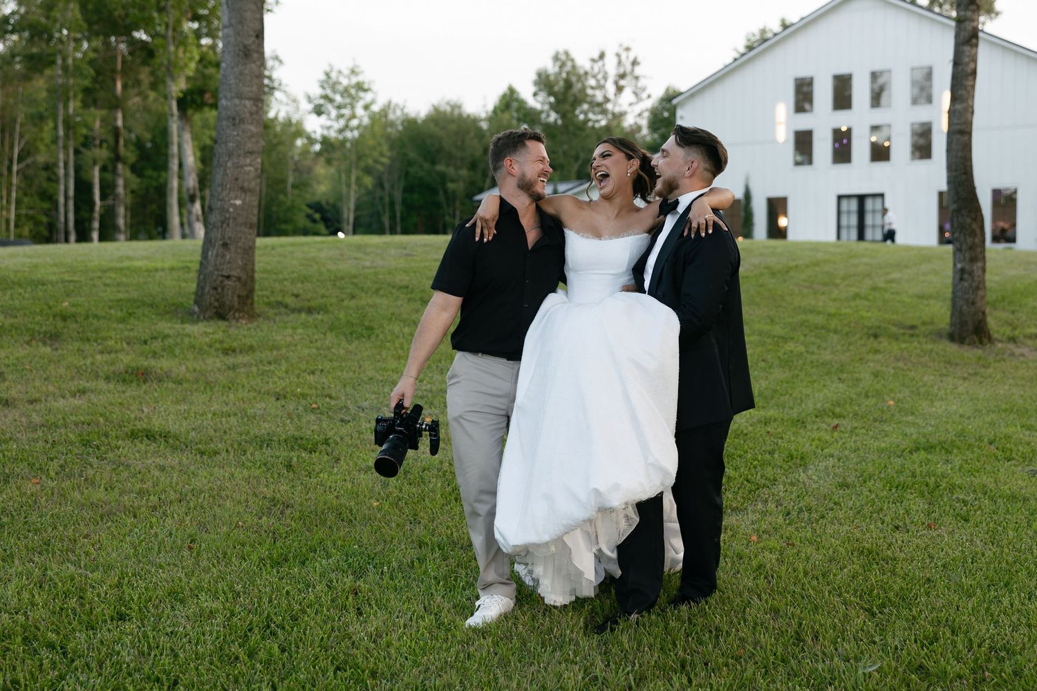 Bride in white gown laughing with two men in black shirts on green lawn near white building and trees.