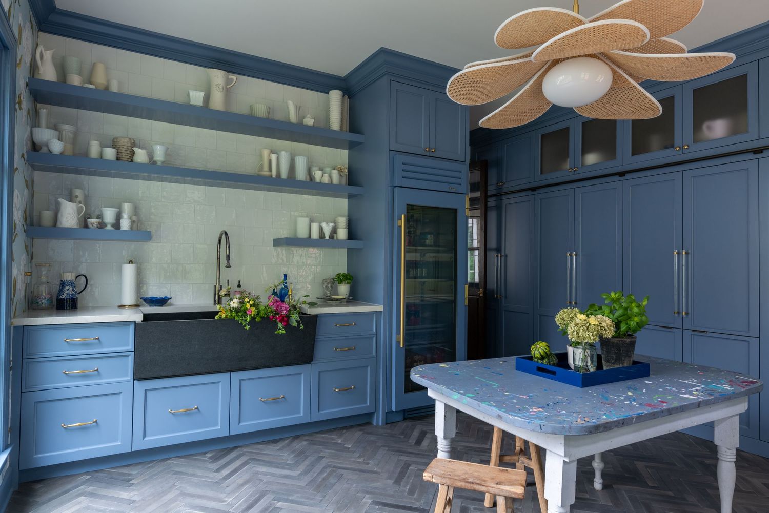 Blue kitchen with herringbone floors, open shelving, farmhouse sink, and a floral pendant light above a marble table.