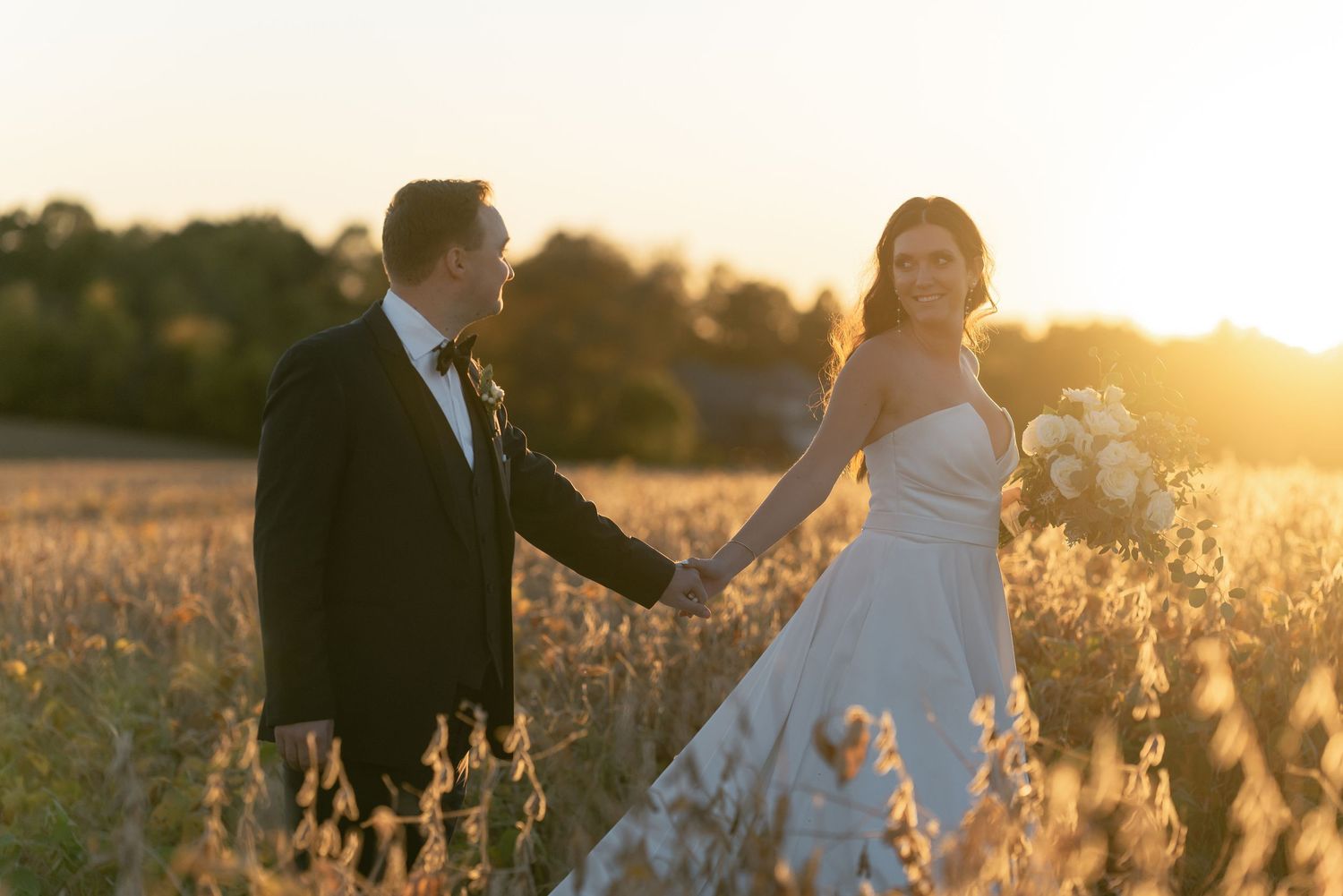 A couple holds hands in a field of golden flowers at sunset while wearing formal wedding attire.