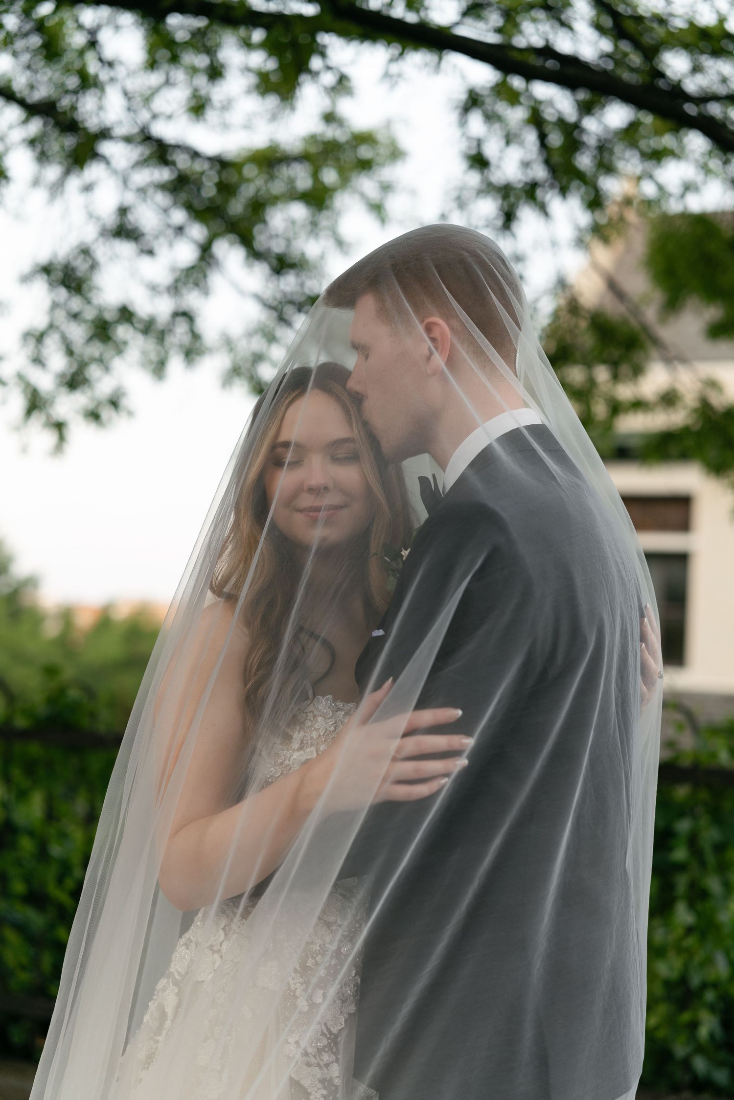A tender moment between newlyweds under a sheer veil in a garden setting.