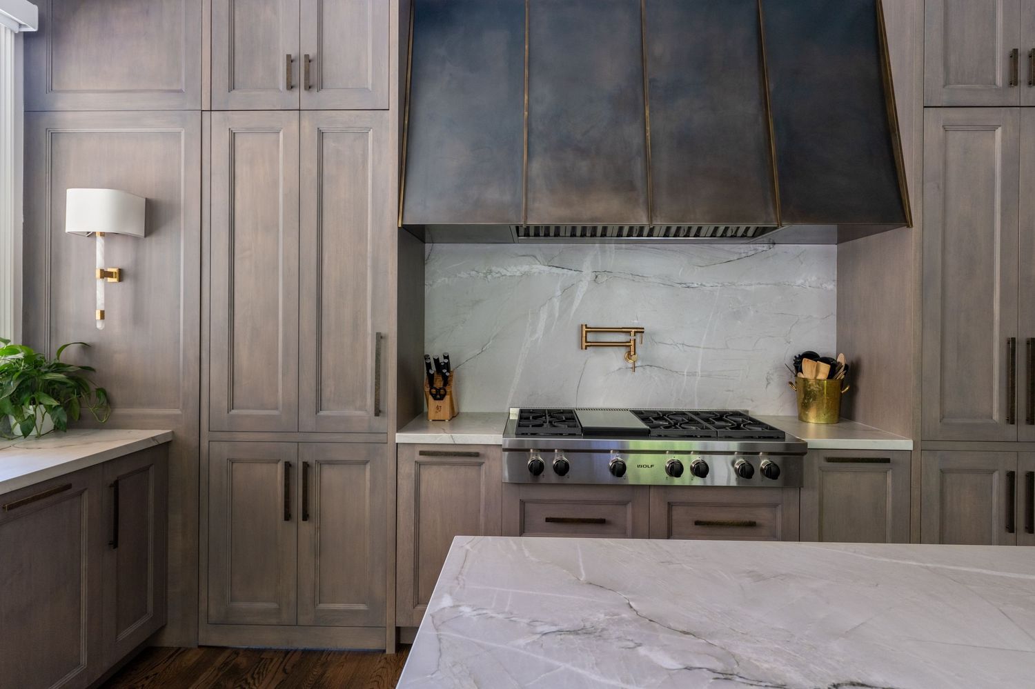Elegant kitchen with gray wood cabinets, marble backsplash, gas range, and a custom metal range hood.