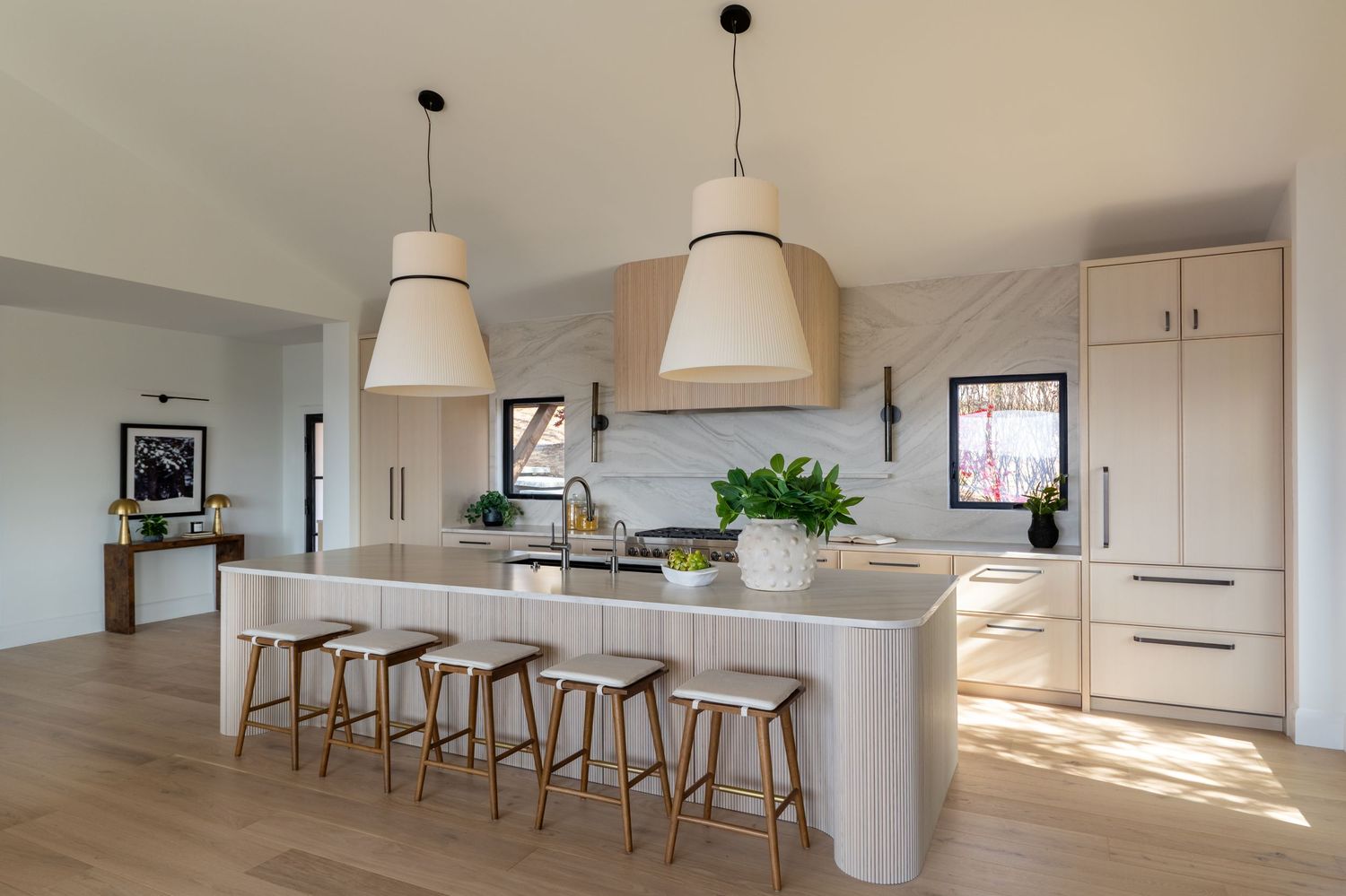 Modern white kitchen with large island, bar stools, pendant lights, and light wood floors in an open-plan layout.