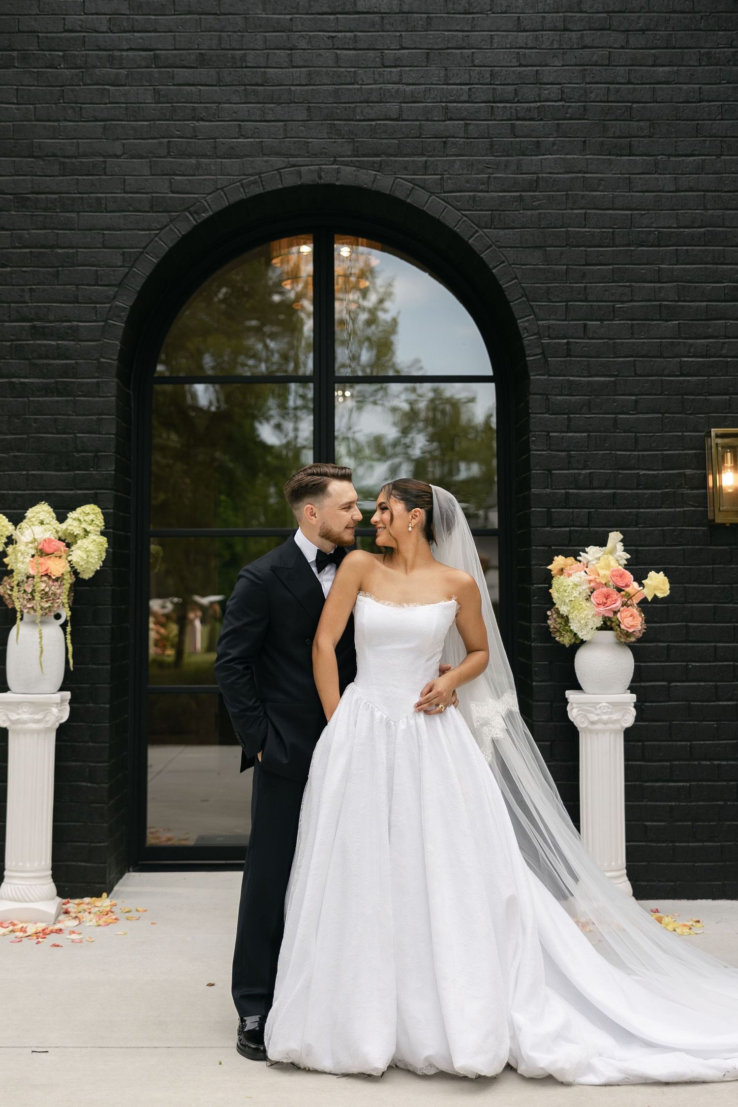 A romantic wedding couple embraces in front of a black brick wall with arched window and floral arrangements.