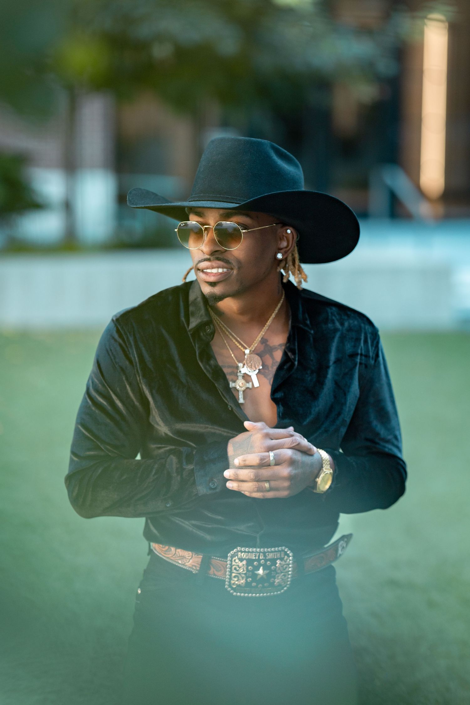 Stylish figure in black cowboy hat and velvet jacket poses outdoors at dusk.