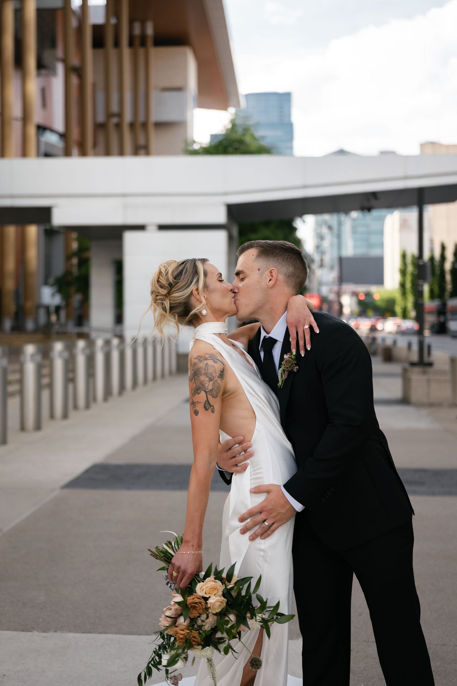 Bride and groom share a kiss outdoors, she holds a bouquet, wearing a backless gown with tattoos visible, he wears a black suit.