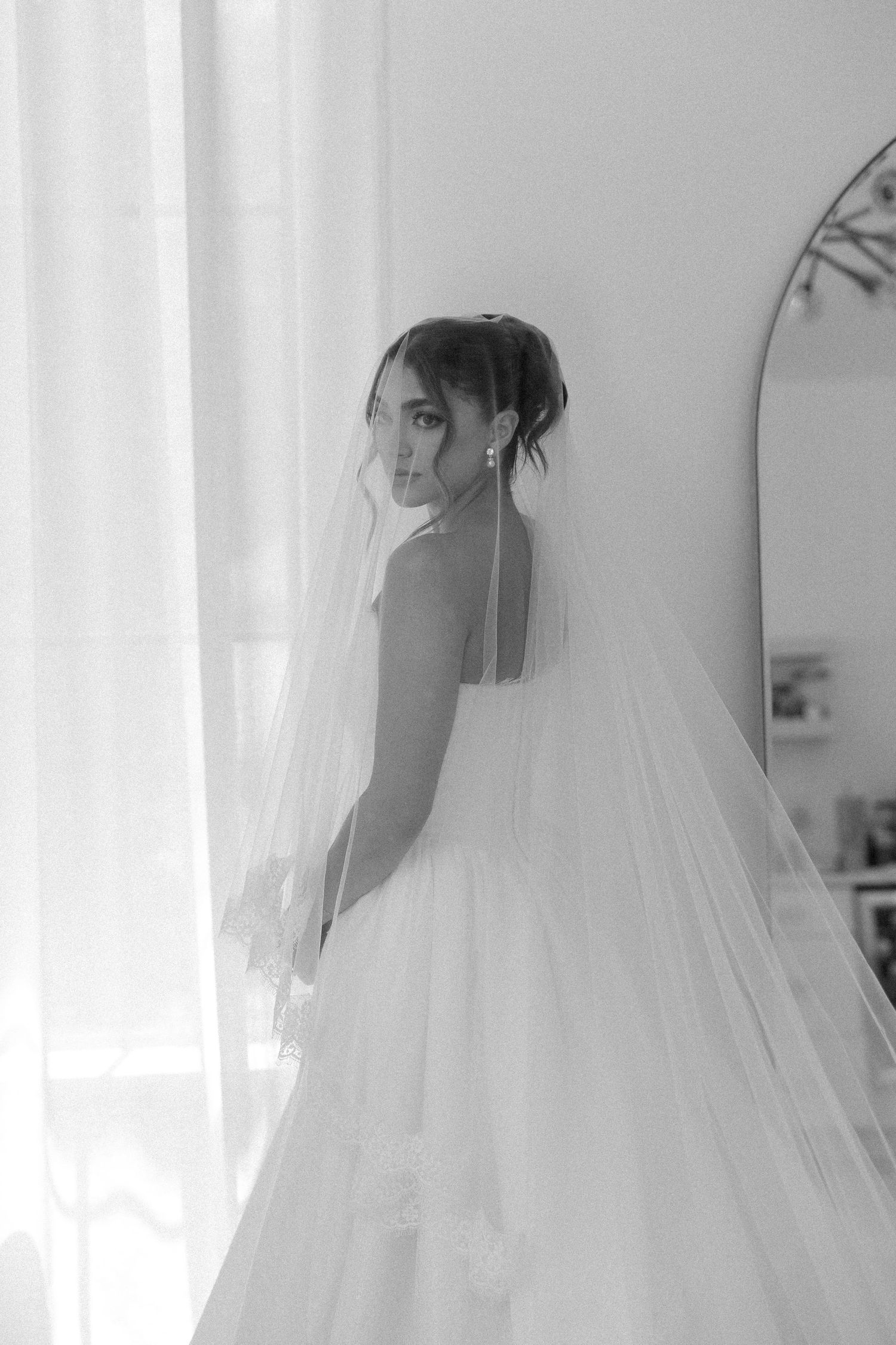 A bride in a flowing white wedding dress and veil stands near a bright window in black and white photography.
