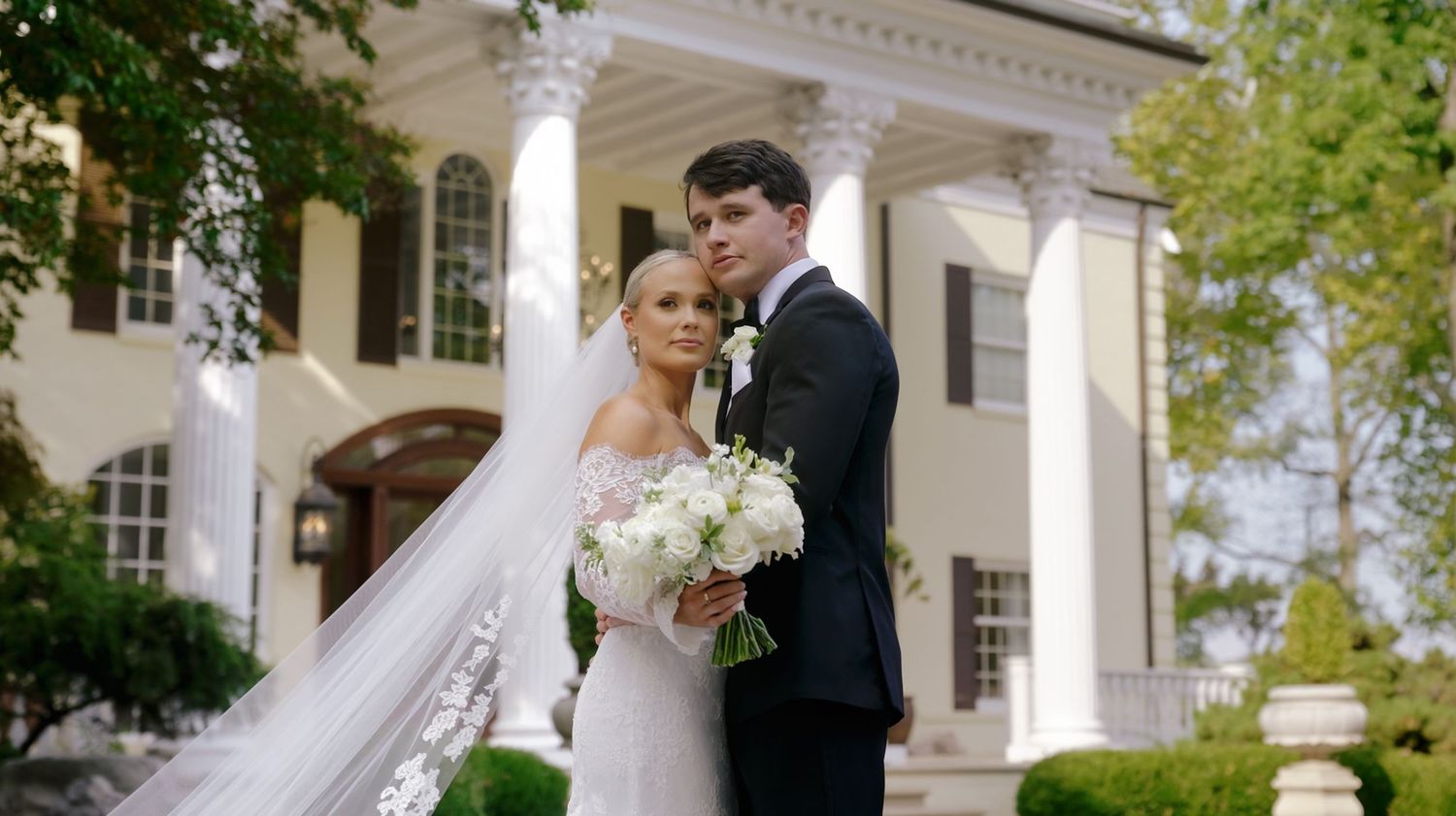 A bride and groom pose in front of a grand white mansion with columns and lush greenery on their wedding day.