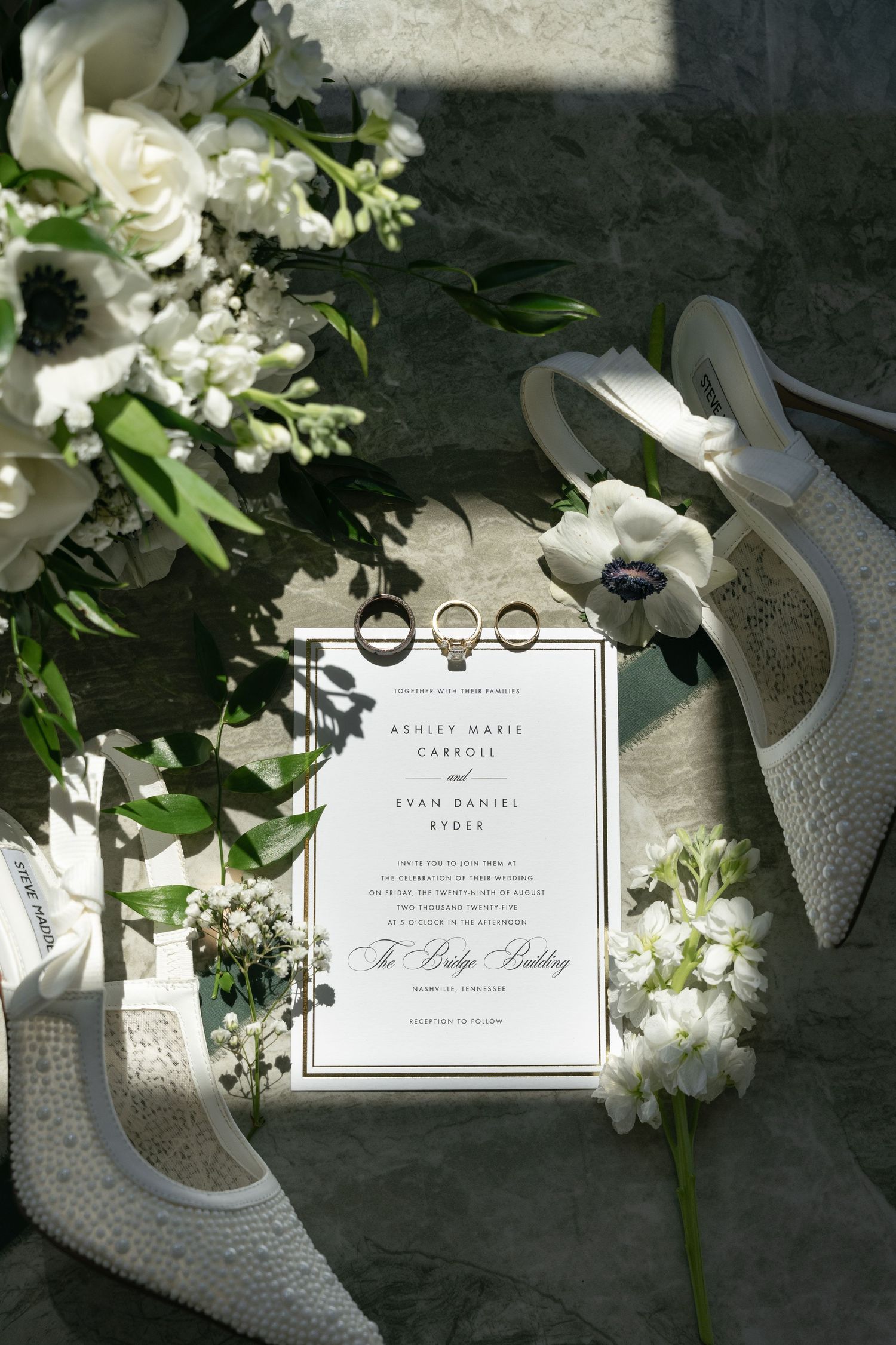 Wedding details featuring white invitation cards, sparkly bridal shoes, and delicate floral arrangements on a textured surface.