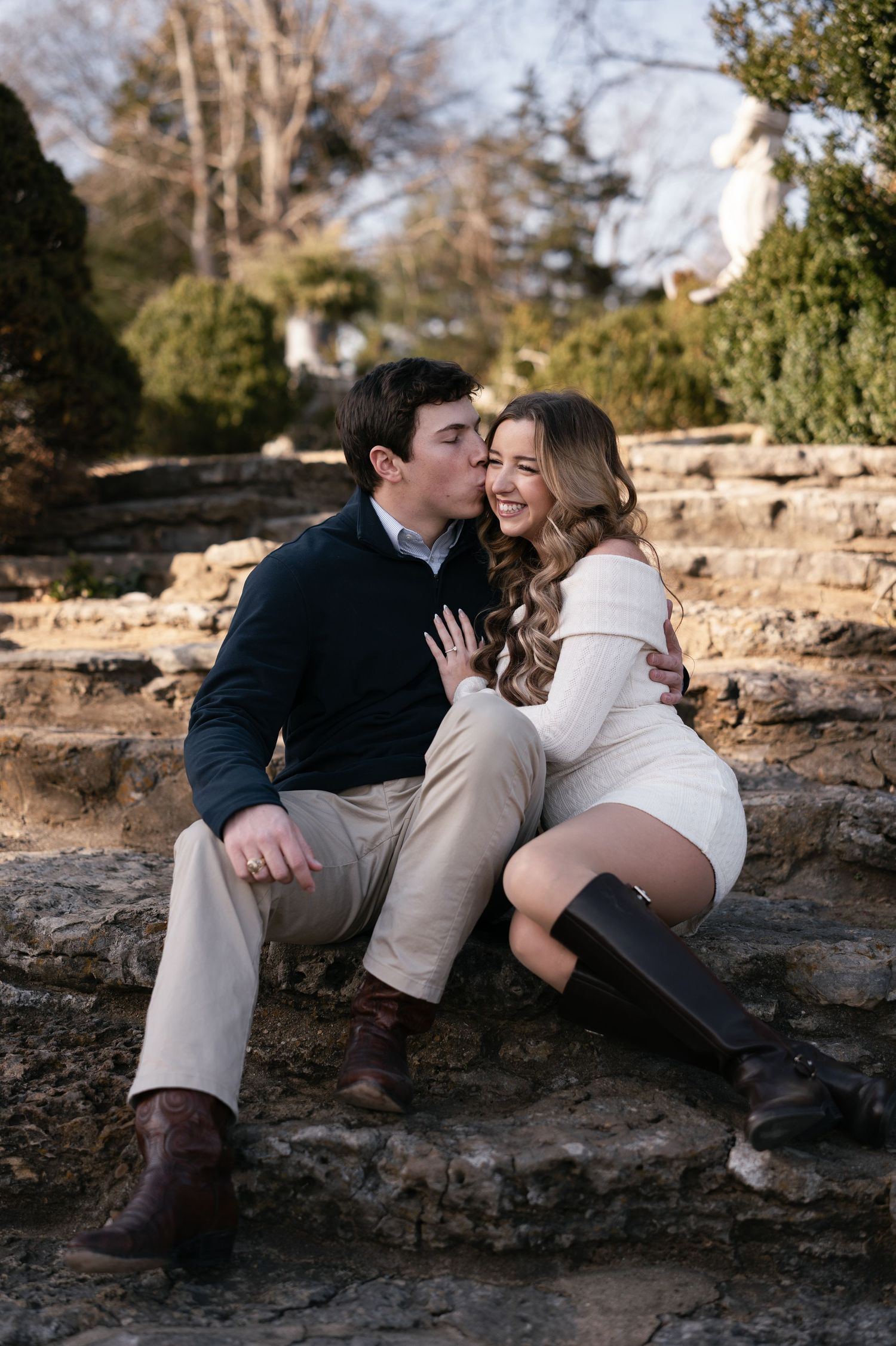 A romantic couple shares tender moments together while sitting on rocky terrain during a sunny outdoor photo session.