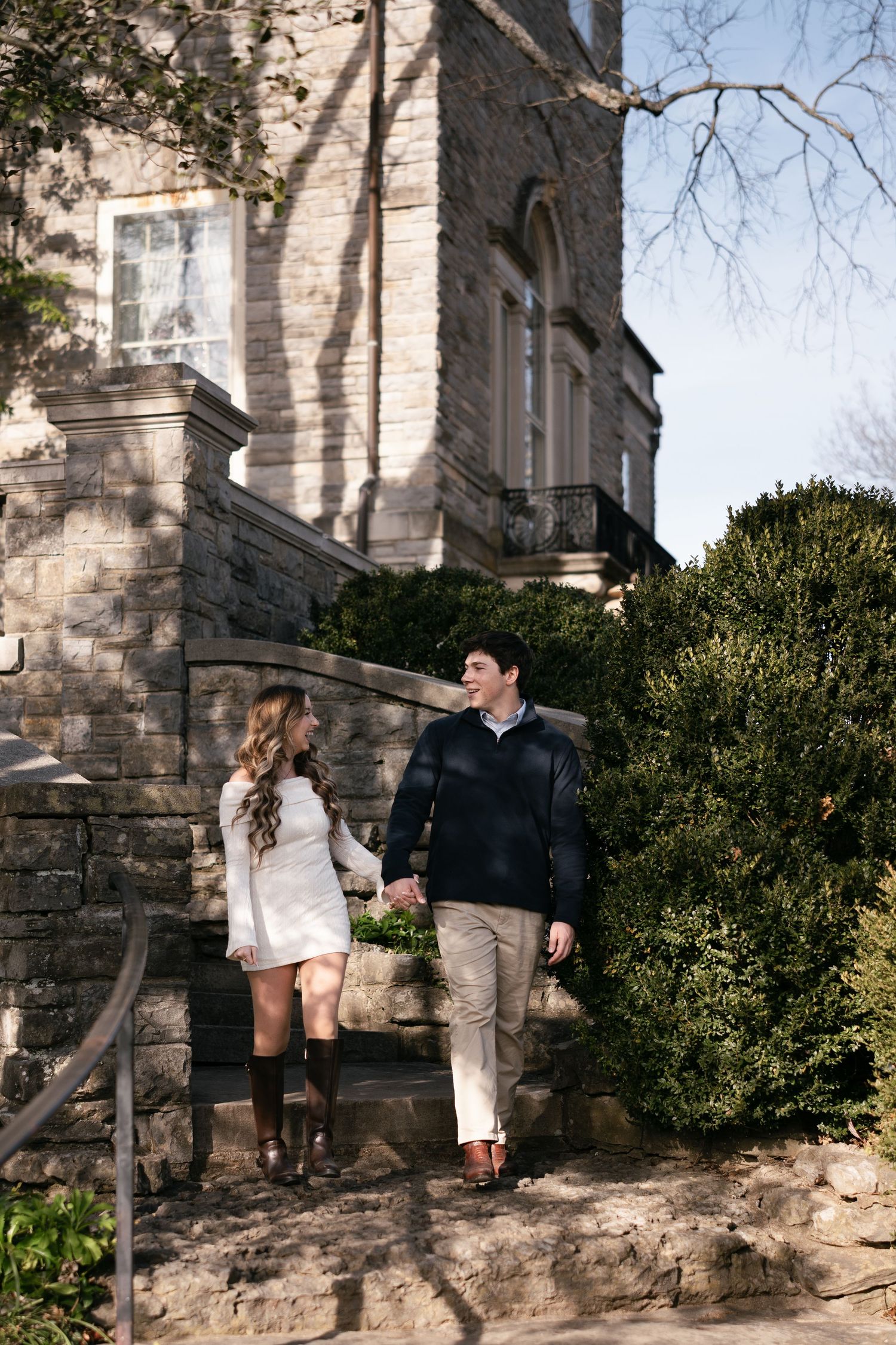 A couple in winter attire shares a romantic moment against a stone building exterior with greenery.