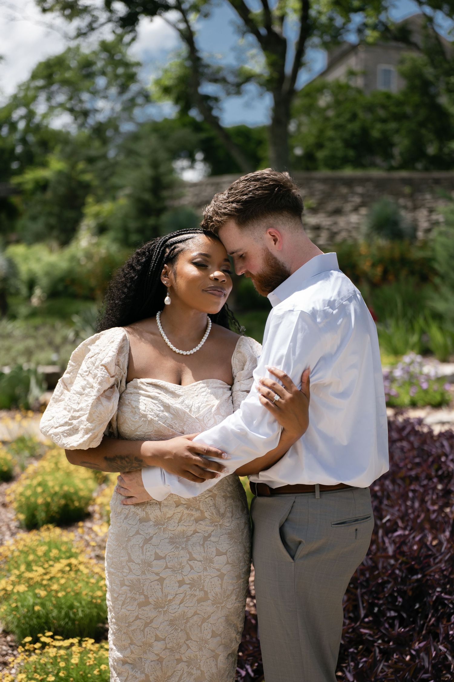 Interracial couple in wedding attire embracing in a lush garden, bride in lace gown with pearls, groom in white shirt.