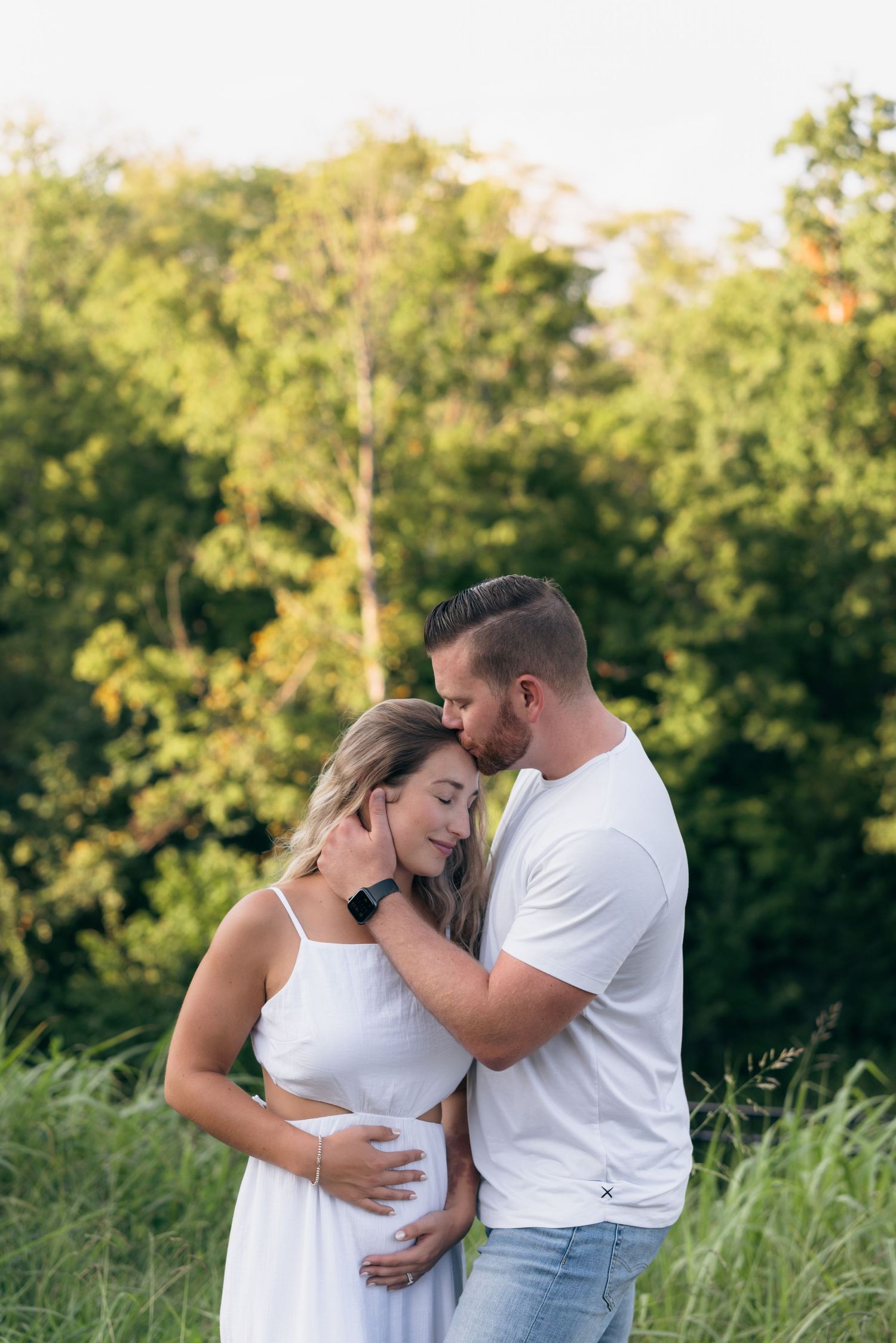 Couple embraces tenderly in a sunlit meadow with lush green trees in the background during golden hour.