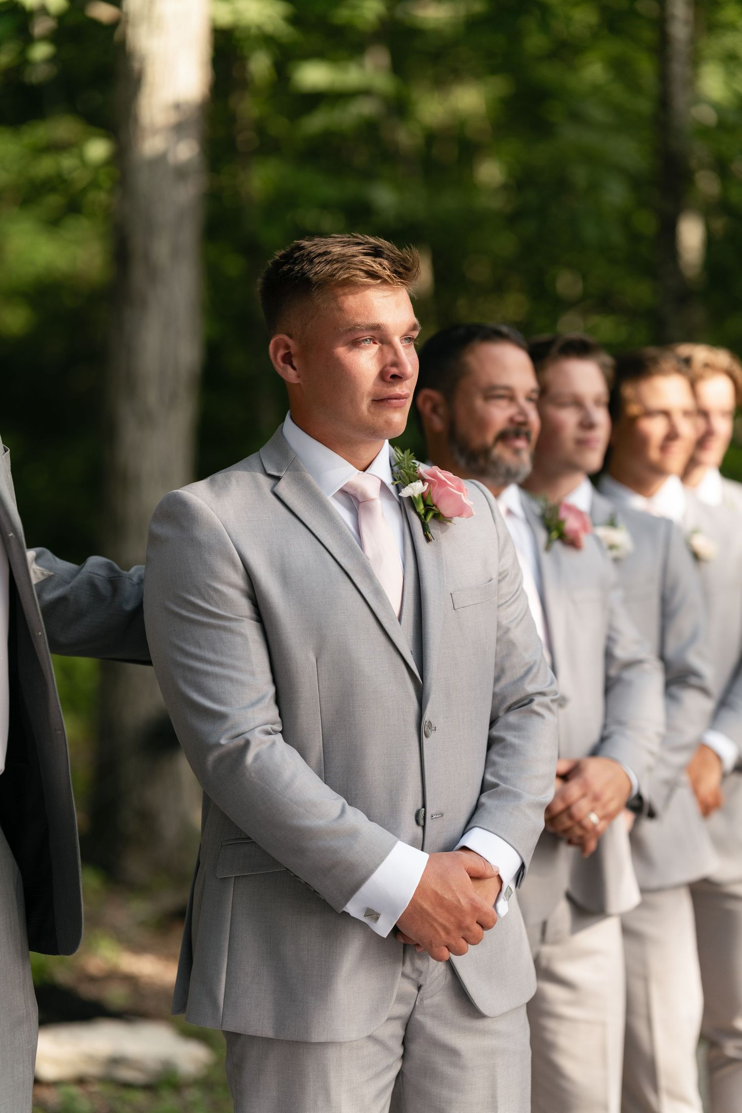 Groomsmen stand in formation wearing matching light gray suits at outdoor ceremony.