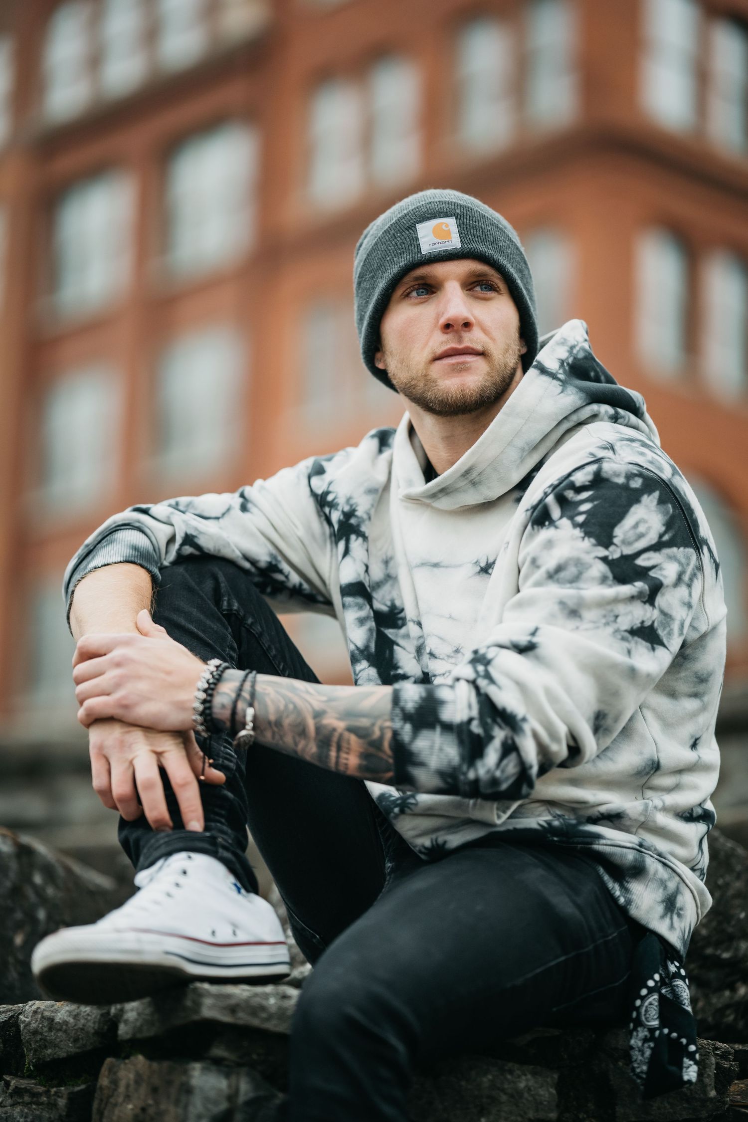 Casual street style portrait in tie-dye hoodie and beanie against brick building background.