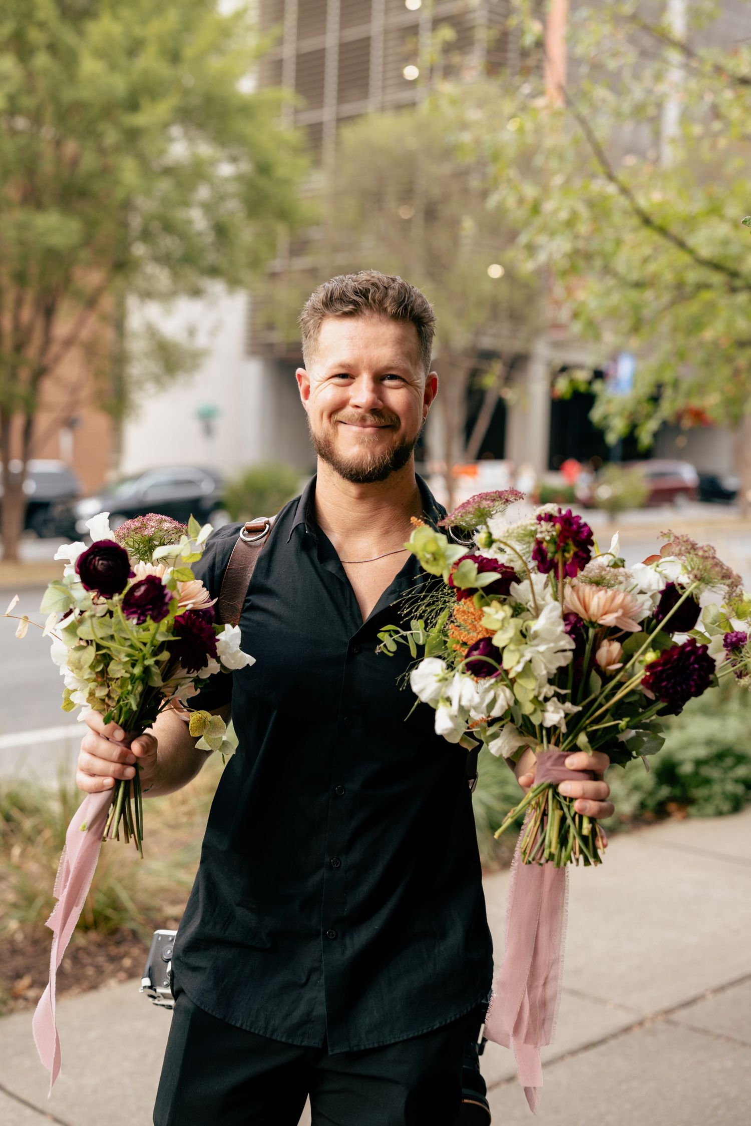 Smiling man in black shirt holding two lush floral bouquets with pink ribbons on a city sidewalk.
