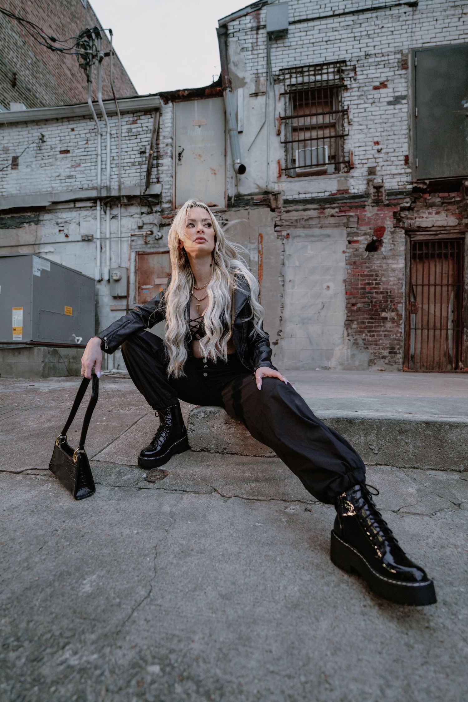 Fashion model in black outfit and combat boots poses against industrial brick wall backdrop.