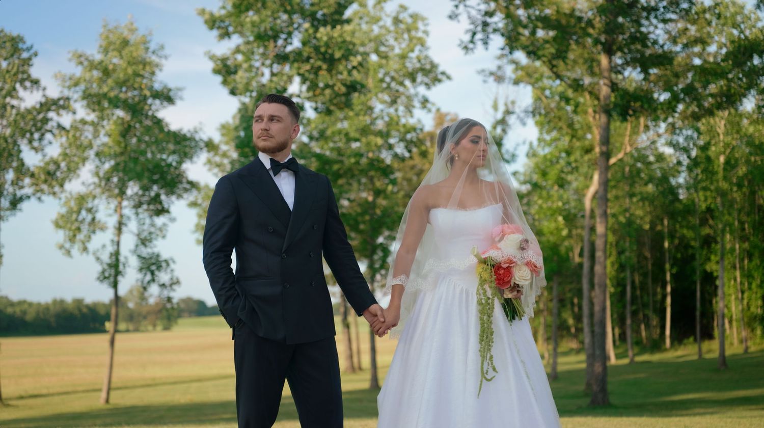 Two people in formal wedding attire walk hand in hand through a lush green outdoor setting at sunset.