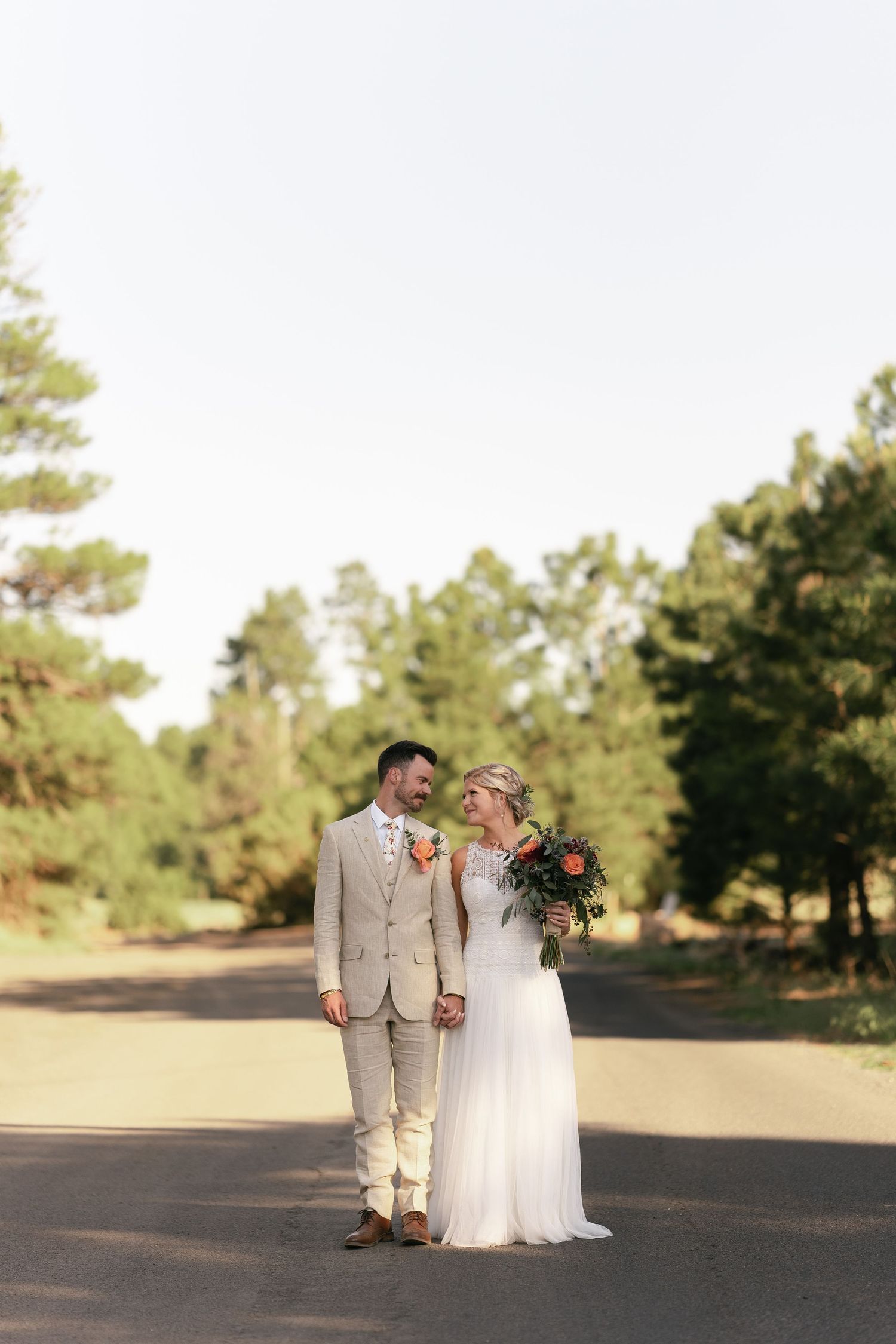 Wedding couple in light-colored attire standing on tree-lined path during outdoor celebration.