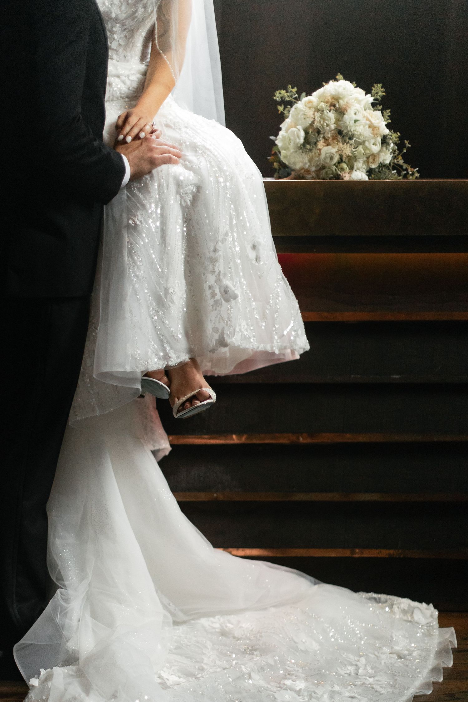 Close-up of a bride's white lace wedding dress and bouquet on wooden stairs with elegant trailing train.