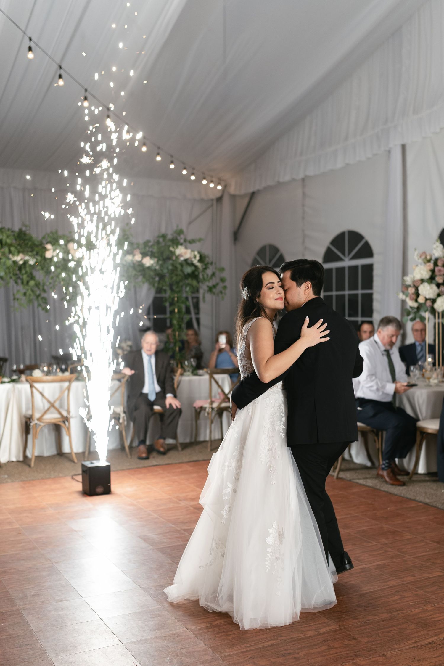A couple shares their first dance at their wedding reception with a dramatic sparkler fountain effect in a white tent.