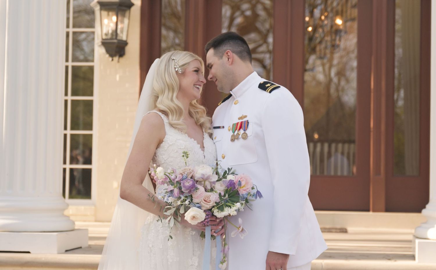 A romantic wedding moment between a bride in white and a military officer in dress uniform holding a purple bouquet.