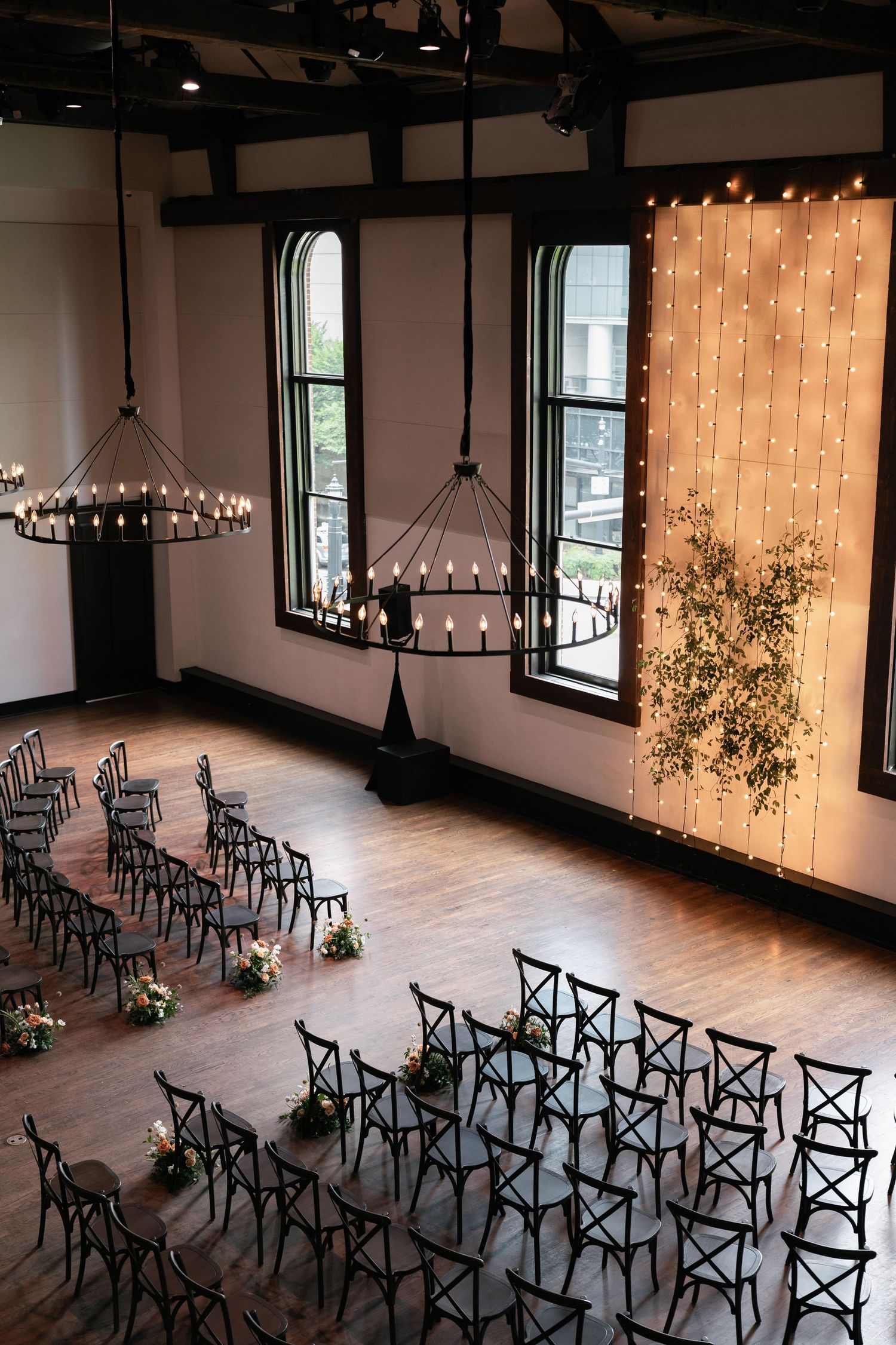 Elegant wedding ceremony venue with black cross-back chairs, fairy light backdrop, greenery, and chandelier lighting on hardwood floors.