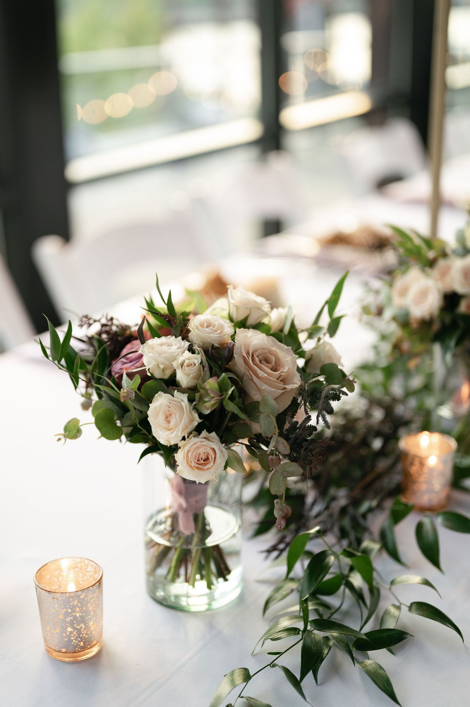 Elegant floral centerpiece with blush roses and greenery in clear vase with mercury glass candle holder.