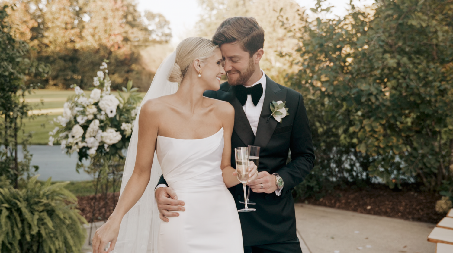 A romantic wedding couple embraces while walking through a garden setting in elegant formal attire.