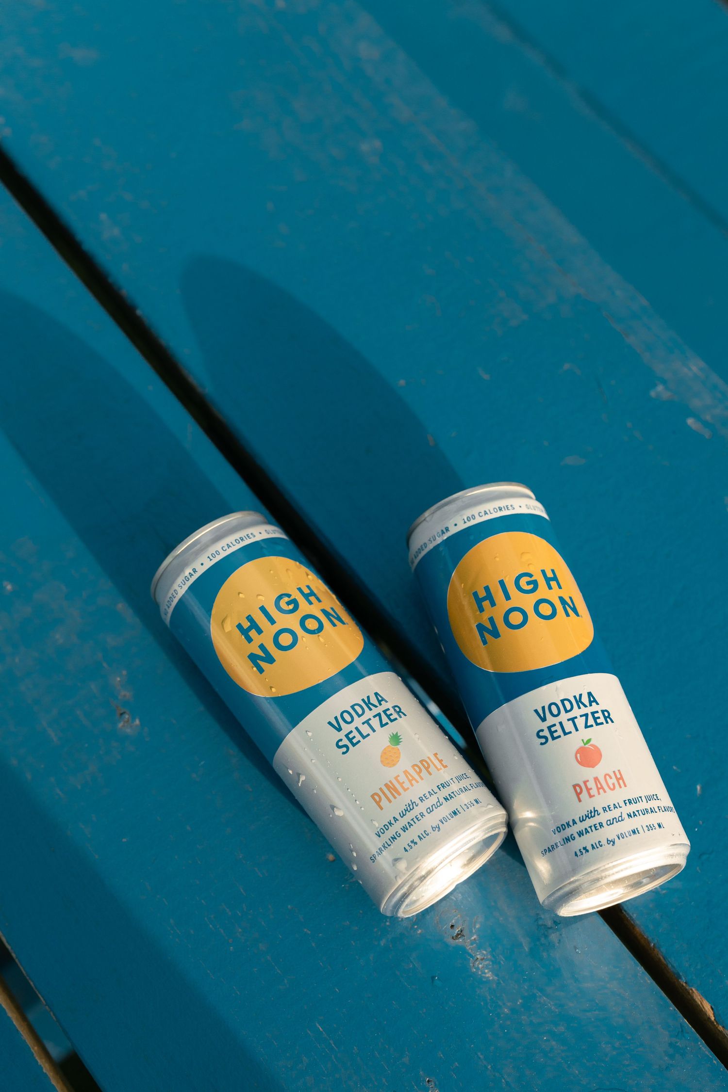 Two High Noon Vodka Seltzer cans in Pineapple and Peach flavors resting on a blue wooden surface.