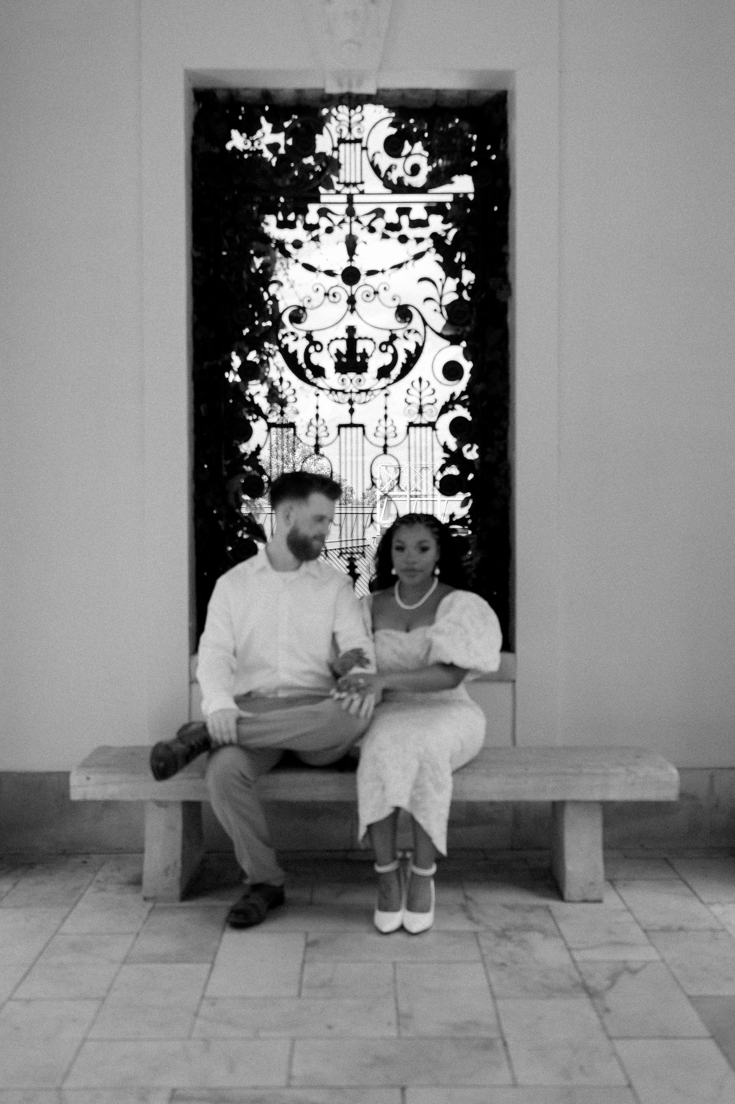 Black and white photo of a couple sitting on a bench in front of a decorative artwork panel.