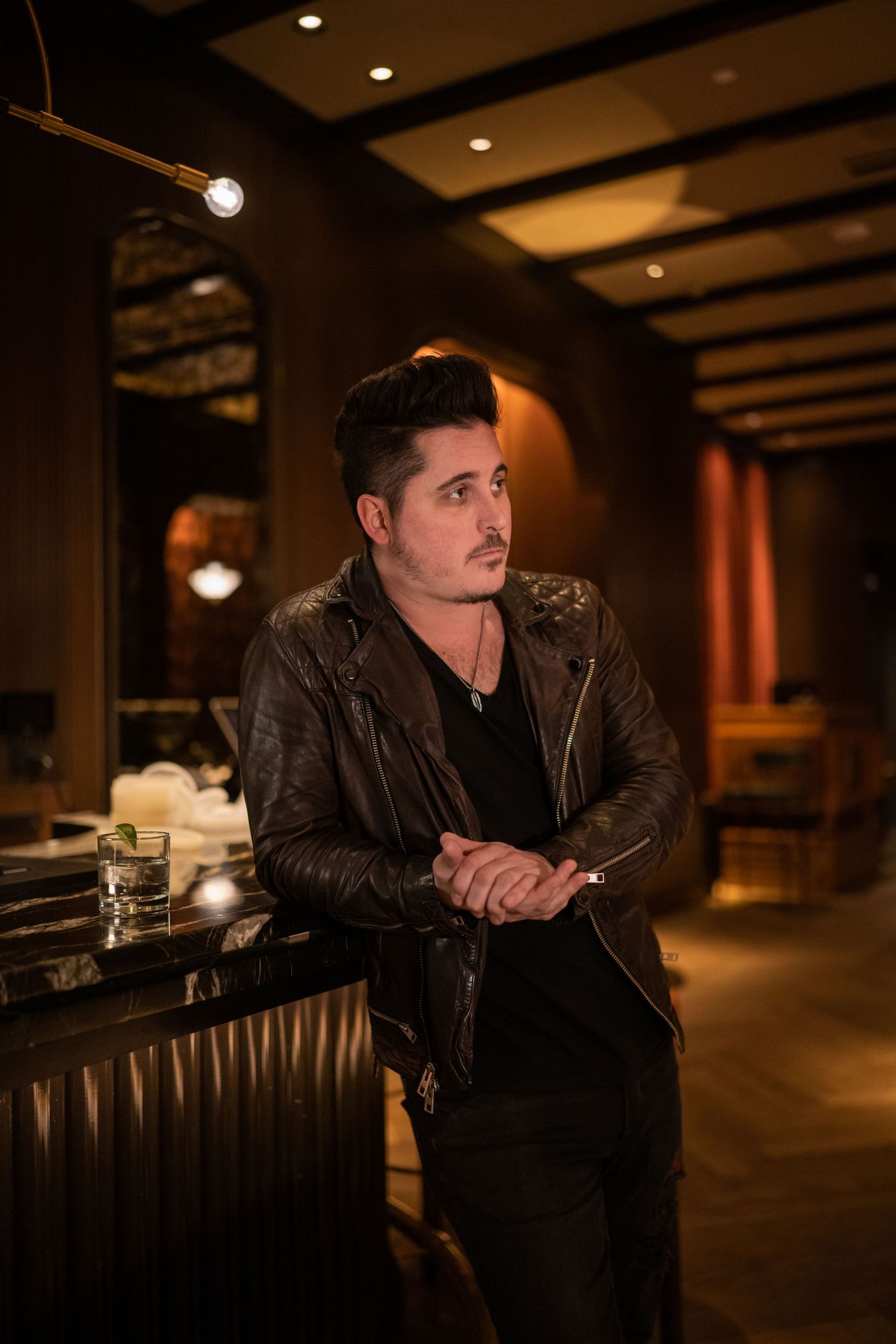Man in leather jacket leans against bar counter in dimly lit restaurant interior.