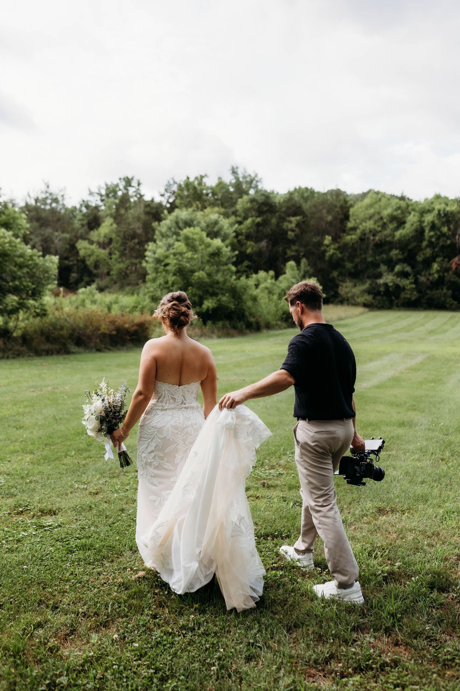 A wedding photographer holds the bride's lace train as they walk across a green lawn surrounded by trees.