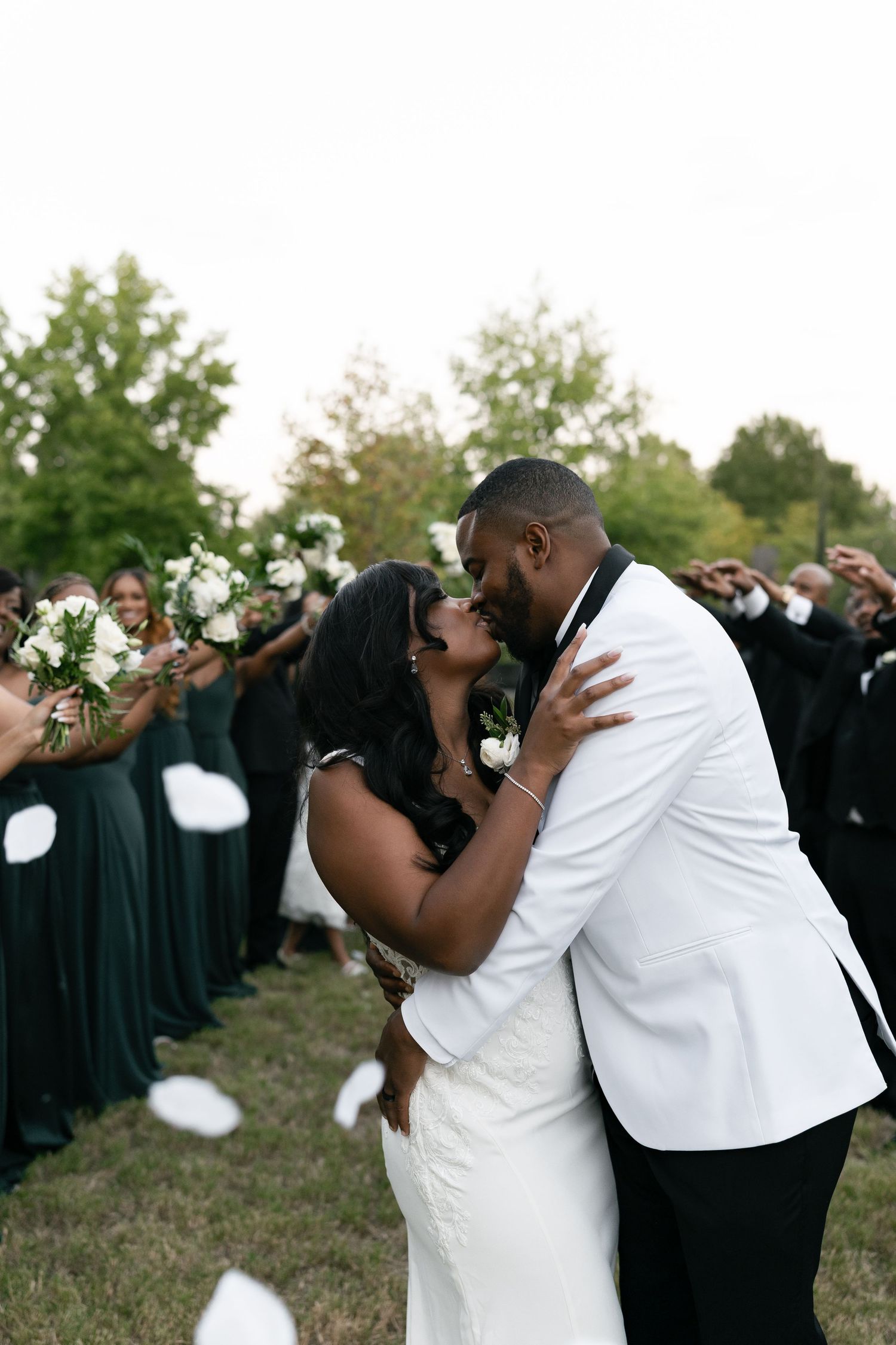 Newlyweds share a romantic kiss surrounded by guests in emerald green dresses throwing flower petals.