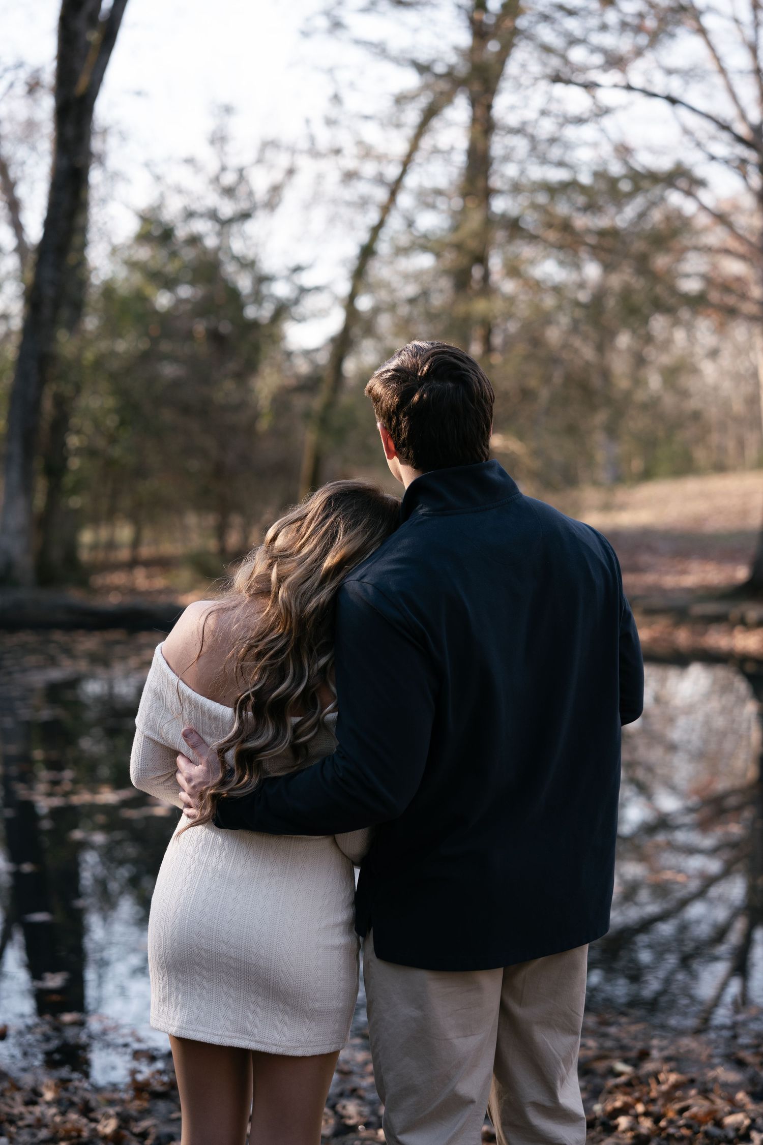 A couple stands together in a romantic embrace against a snowy winter forest backdrop with fallen leaves.
