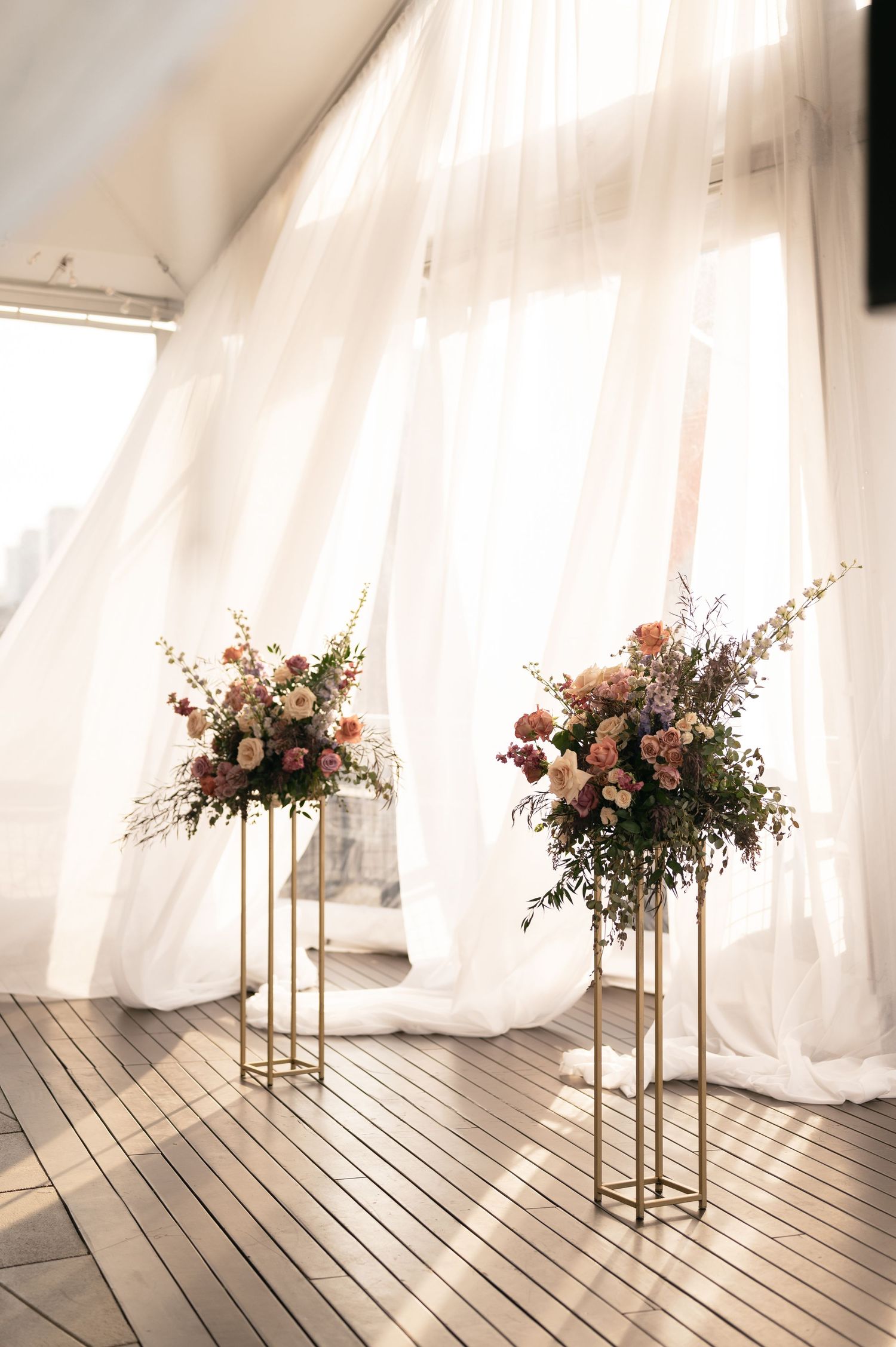 Two tall gold stands with elegant floral arrangements against white flowing curtains in a bright wedding venue setting.