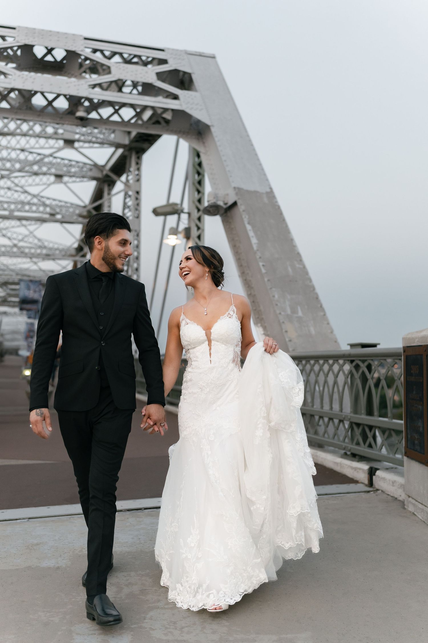 Newlyweds walking hand in hand on bridge while bride's white dress flows in the wind.