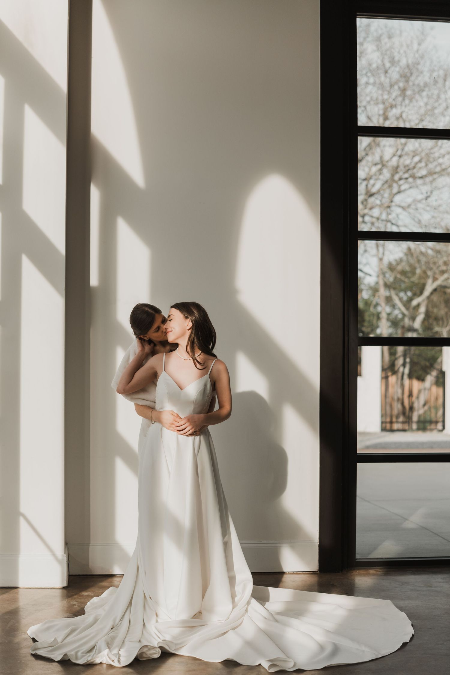 A white wedding gown with a flowing train photographed in a sunlit room with modern windows.