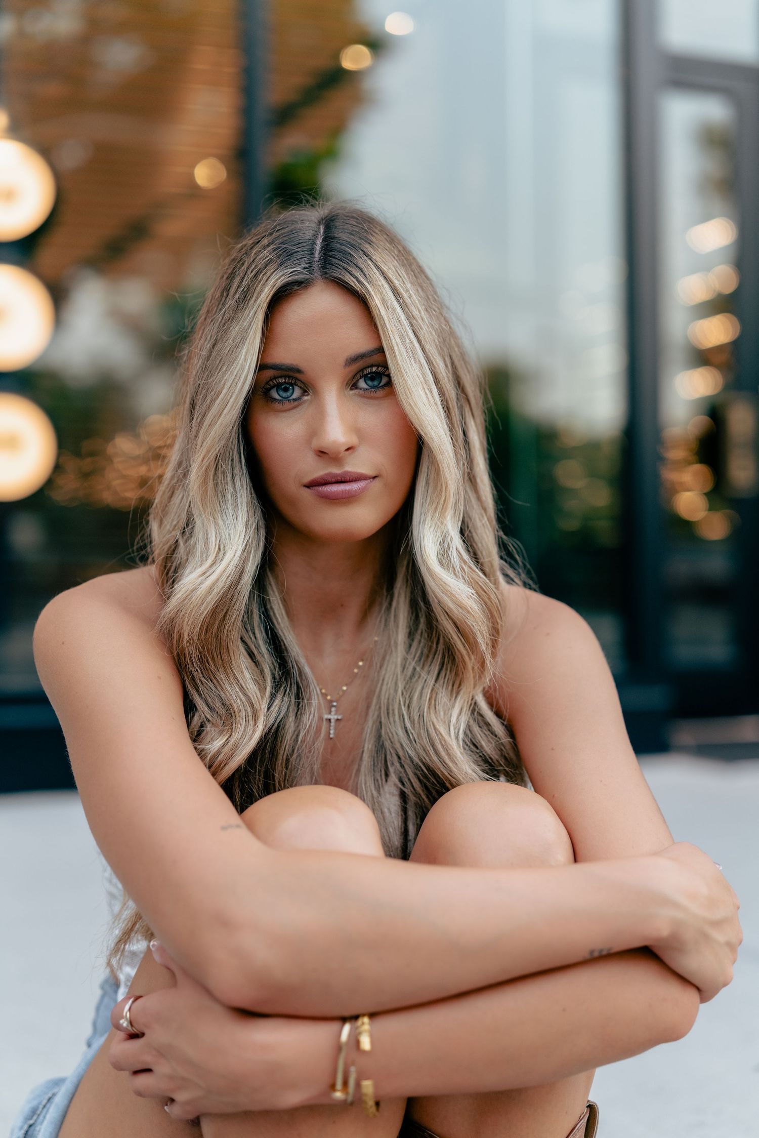 Portrait of woman with wavy hair sitting outdoors with blurred lights in background.
