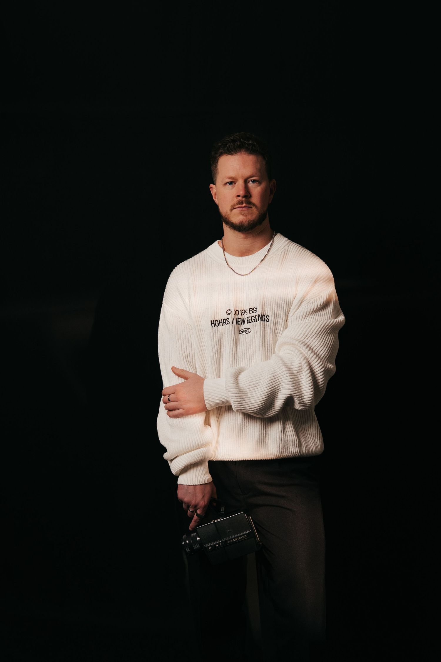 Portrait of person in white knit sweater holding camera against black background.