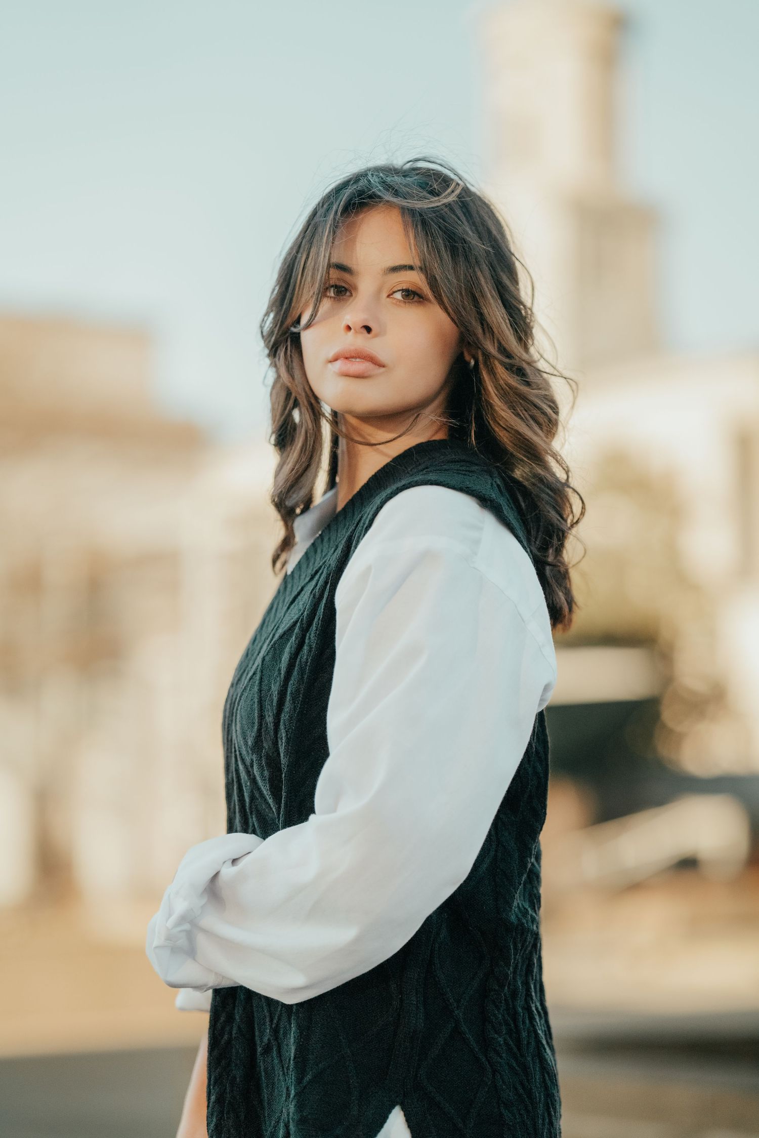 Portrait in a dark knit vest and white blouse against an urban backdrop with warm lighting.