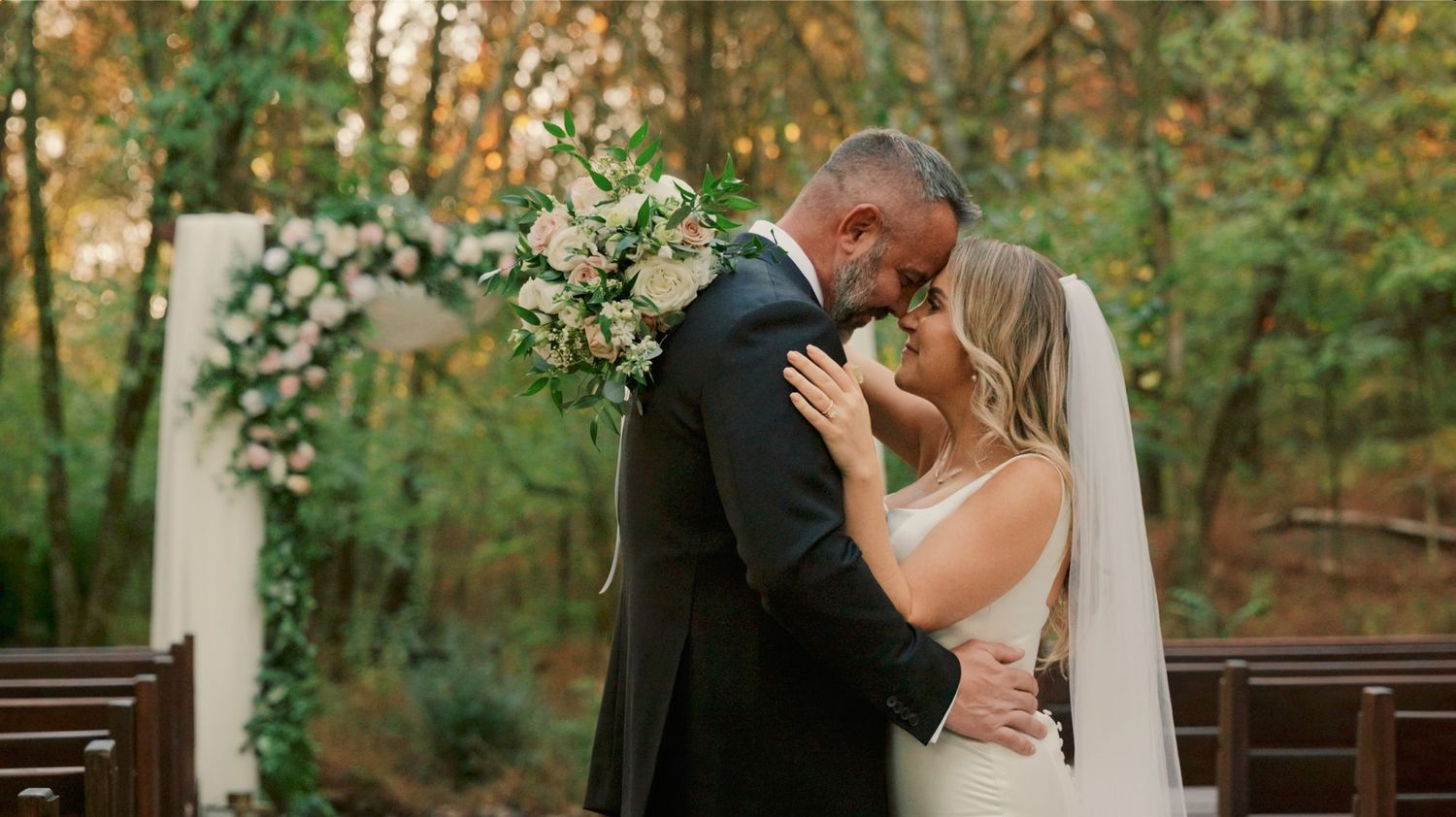 Intimate outdoor wedding ceremony with couple sharing tender embrace under floral arch in wooded setting.