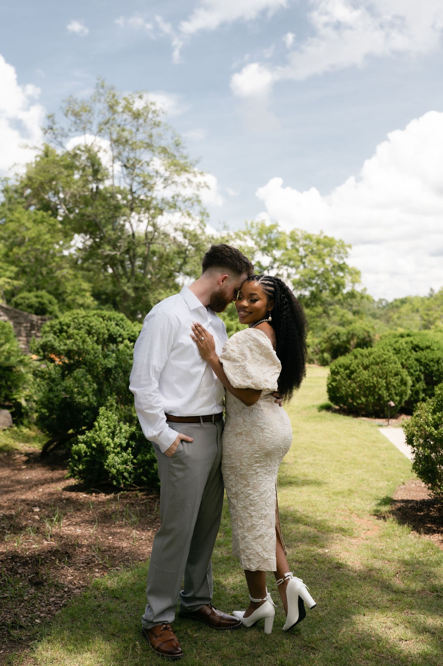 Interracial couple sharing a romantic moment in a lush garden, bride in white lace dress and groom in gray pants.