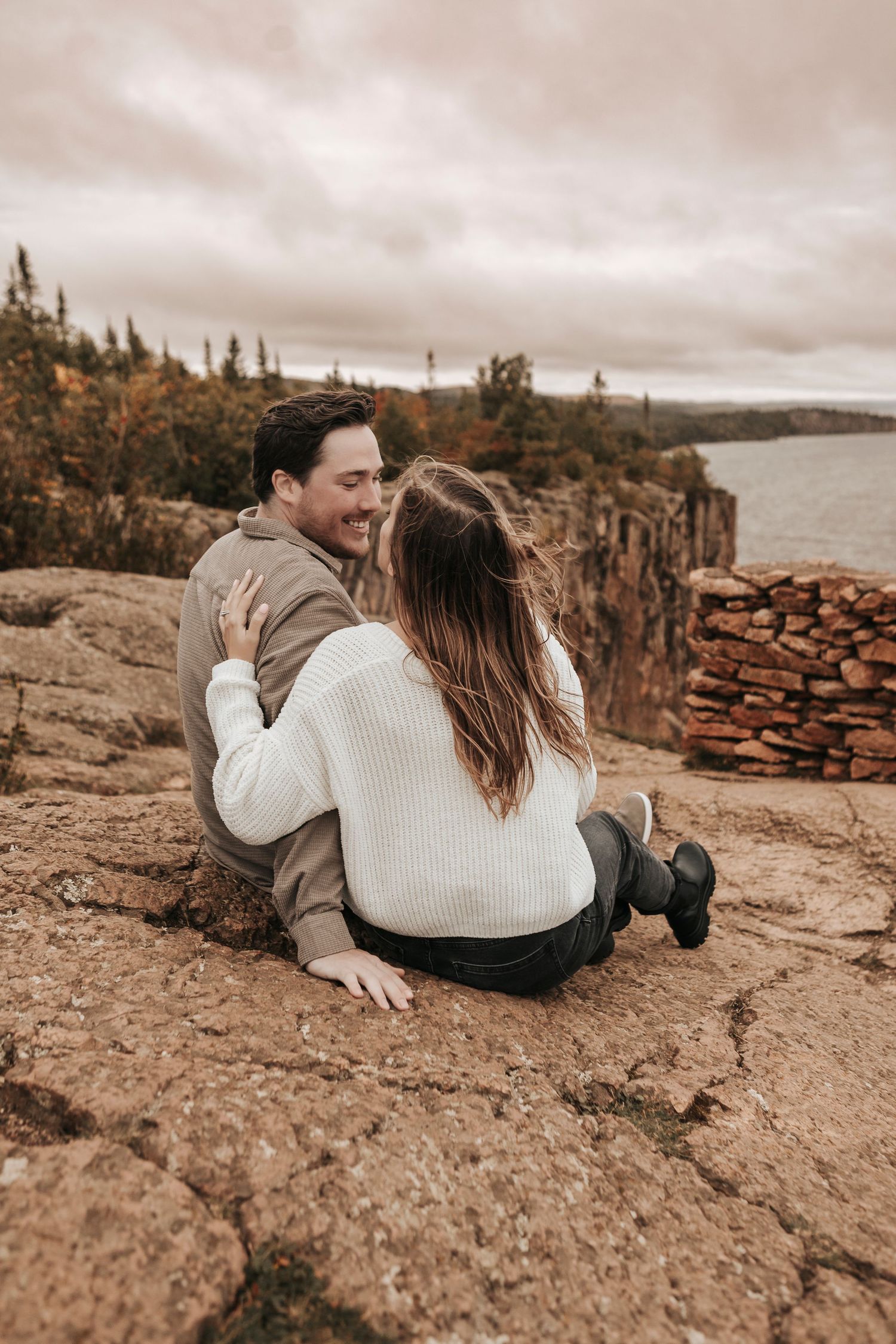 A couple sits together on a rocky cliff overlooking a scenic lake with fall foliage and moody skies in the background.