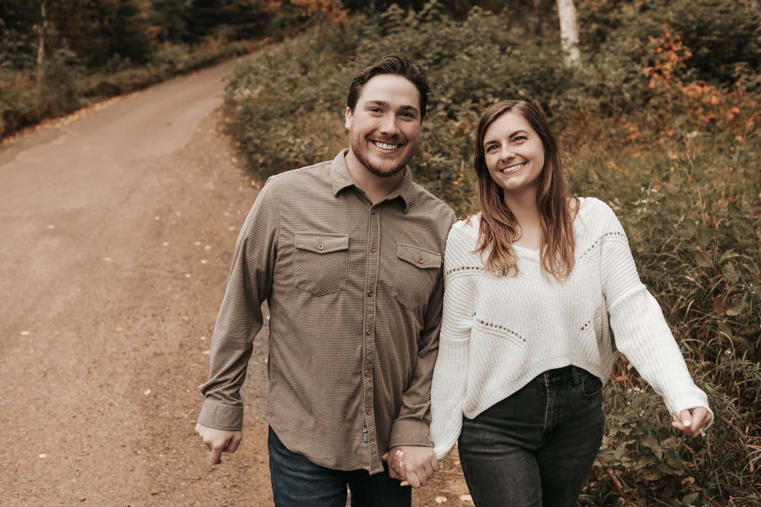 A couple in earth-toned clothing walking and laughing together on a wooded autumn trail.