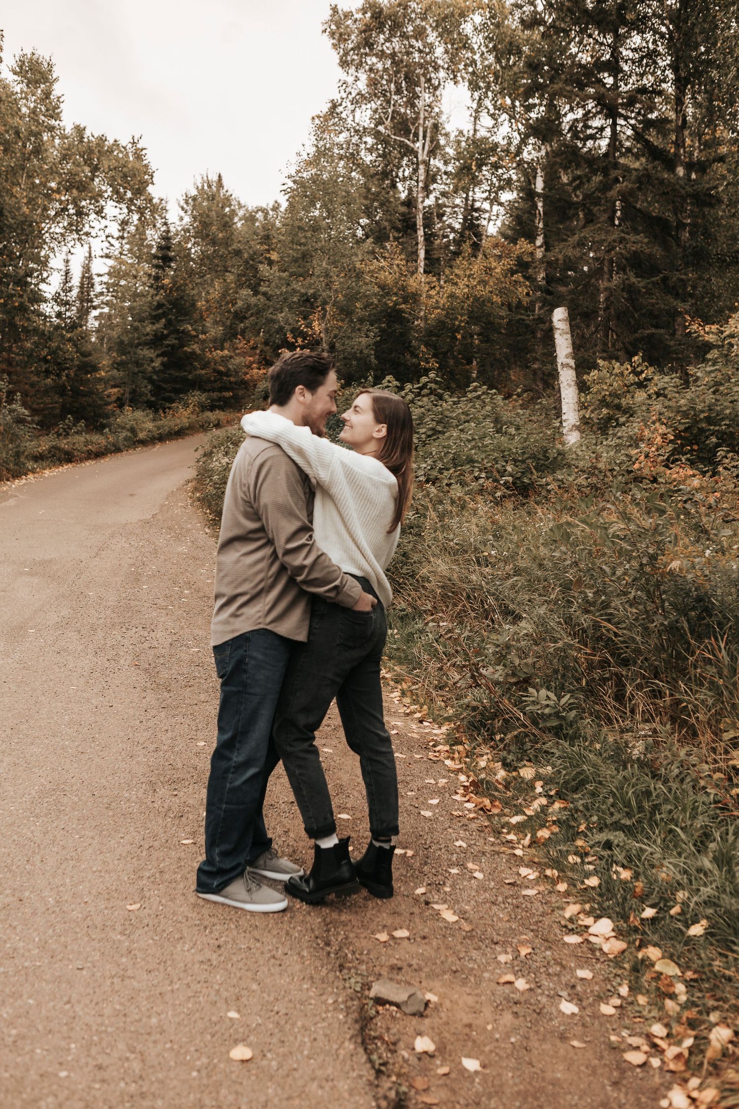 A couple embraces on a winding autumn path surrounded by fallen leaves and trees.