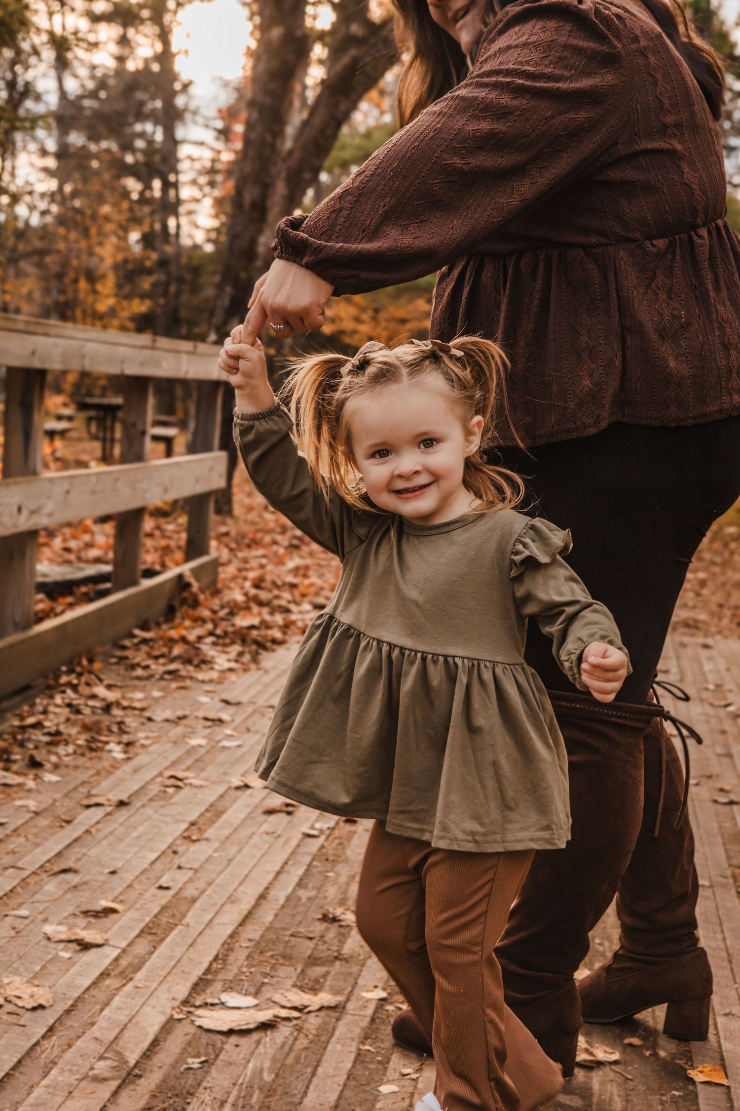 A small child in brown and olive clothing plays on a wooden boardwalk covered in autumn leaves.