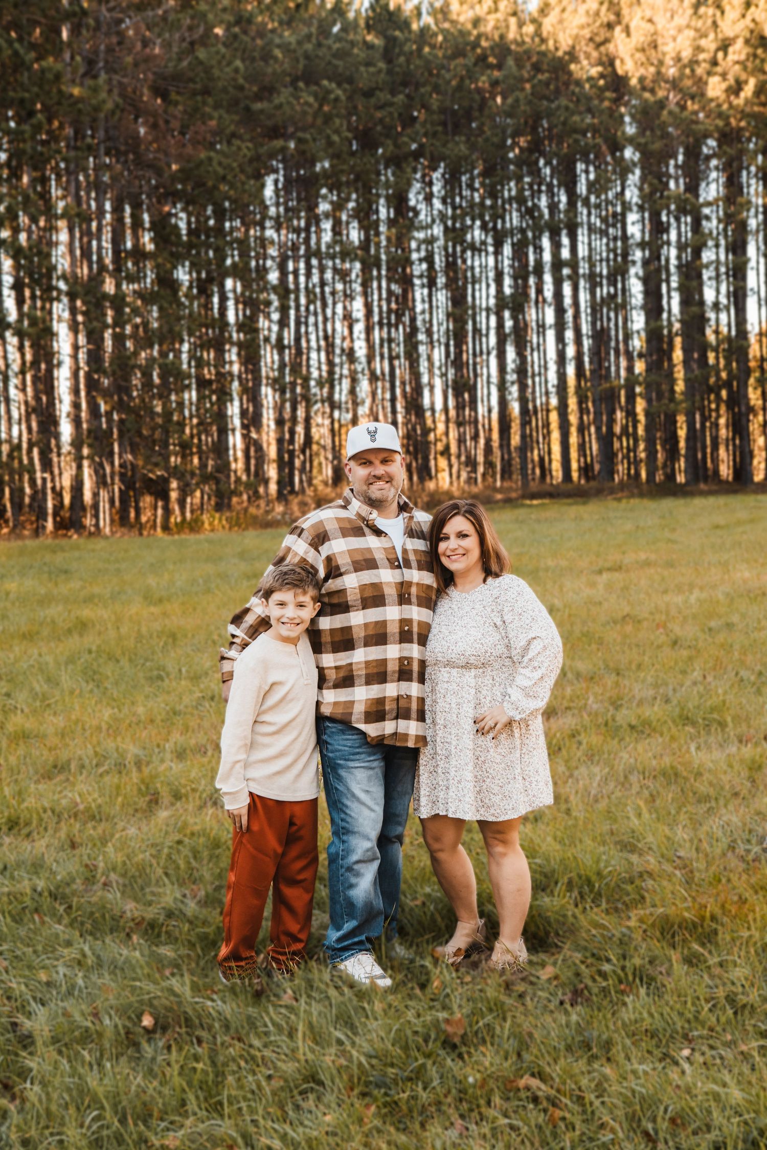 A family stands together in a grassy field at sunset with tall pine trees creating a scenic backdrop.