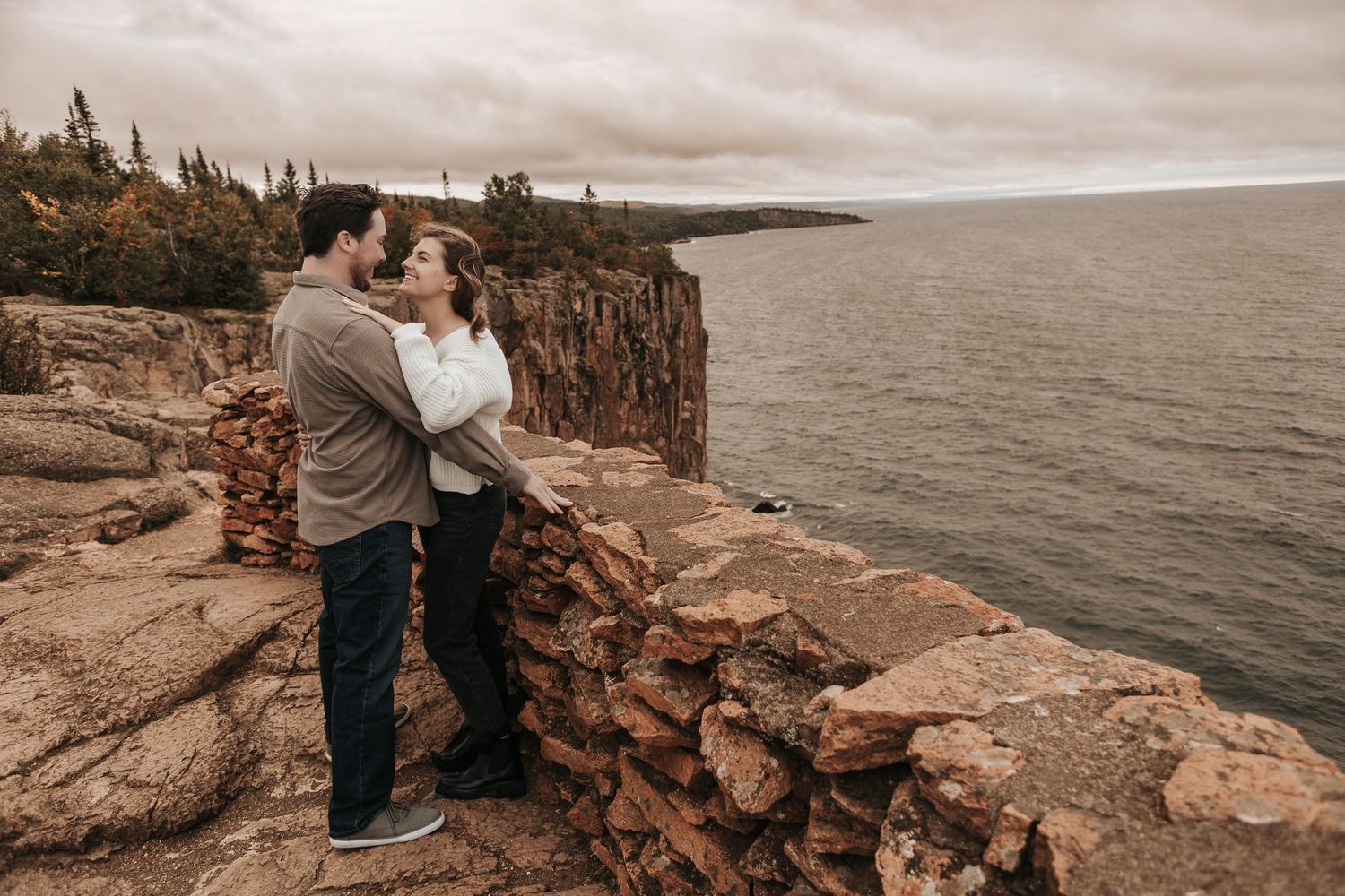 Couple embraces on rocky ocean cliffs during cloudy sunset along rugged coastal shoreline.